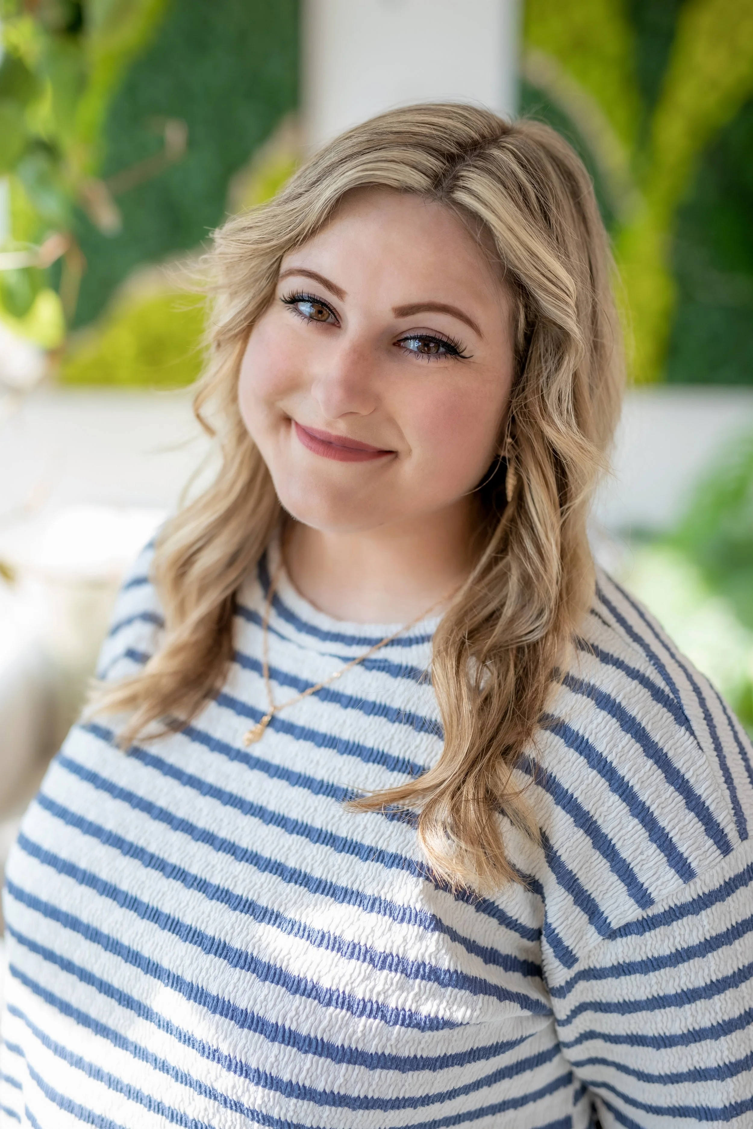 A young woman with wavy blonde hair and brown eyes smiling, wearing a blue and white striped shirt and a gold necklace, with a blurred green outdoor background.