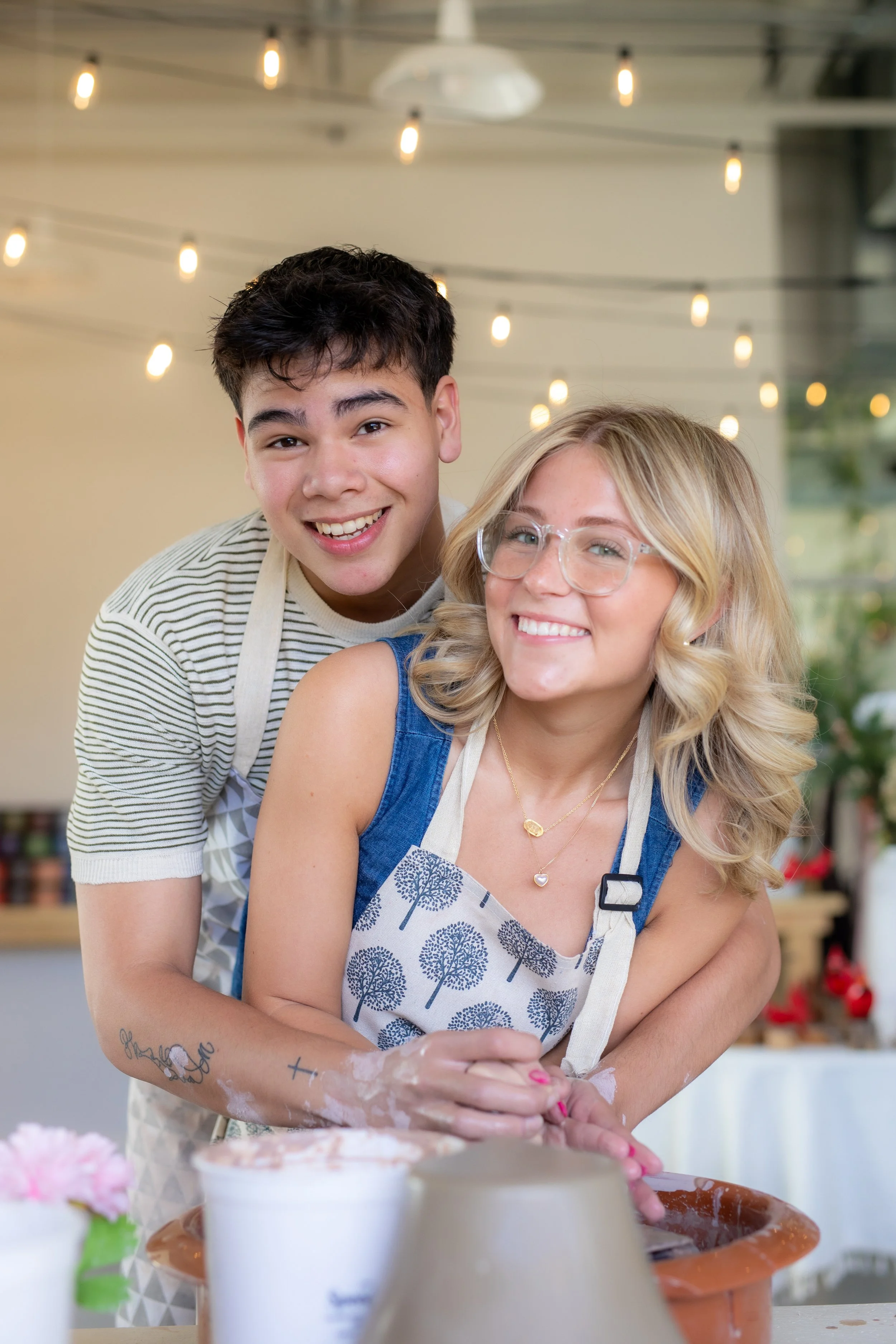 A young man and woman smiling while making pottery with string lights overhead. They are wearing aprons, and their hands are covered in clay as they work on the pottery wheel.