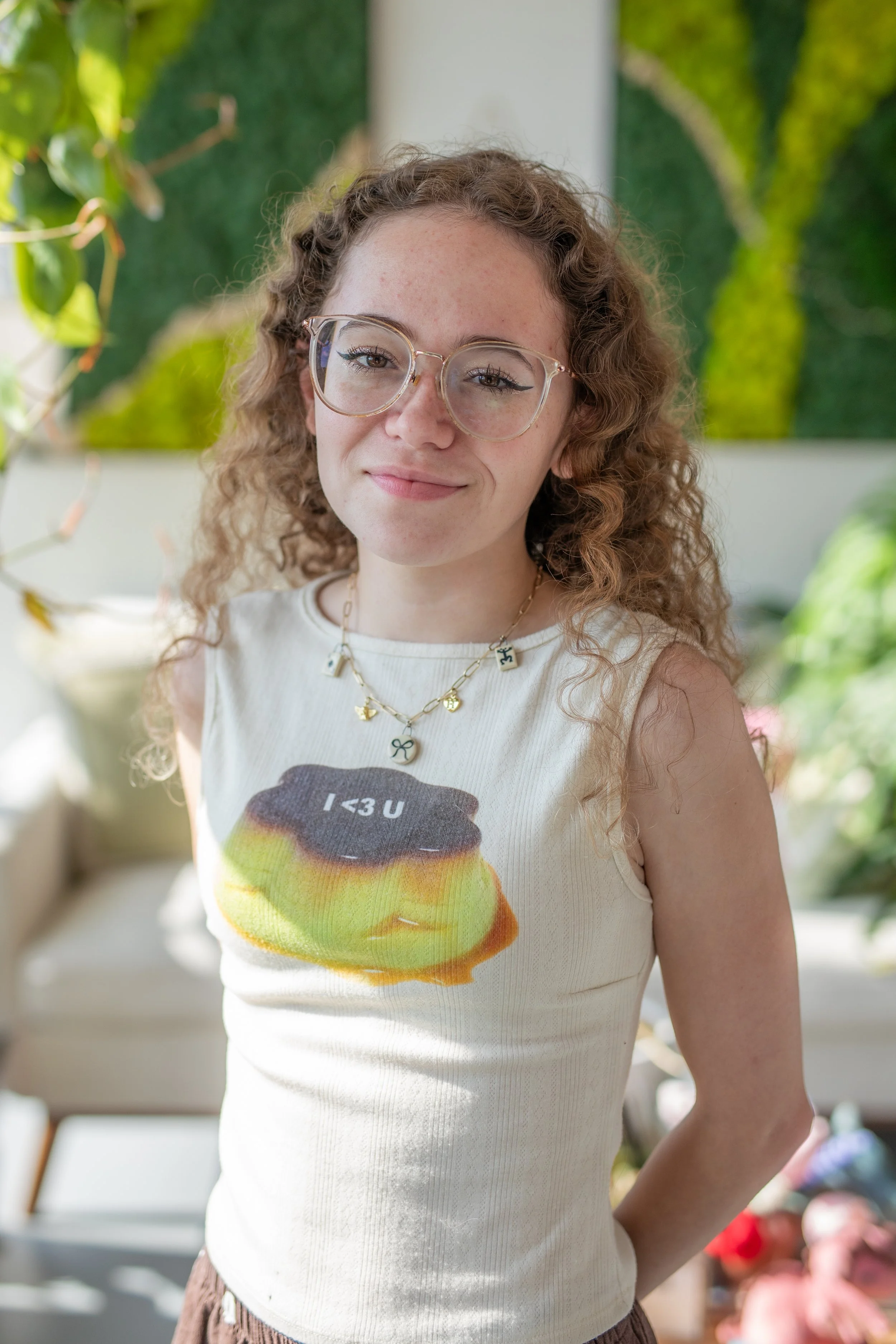 A young woman with curly hair and glasses smiling outdoors, wearing a sleeveless shirt with a rainbow-colored donut graphic and text, and a necklace, in a bright, green, garden-like setting.