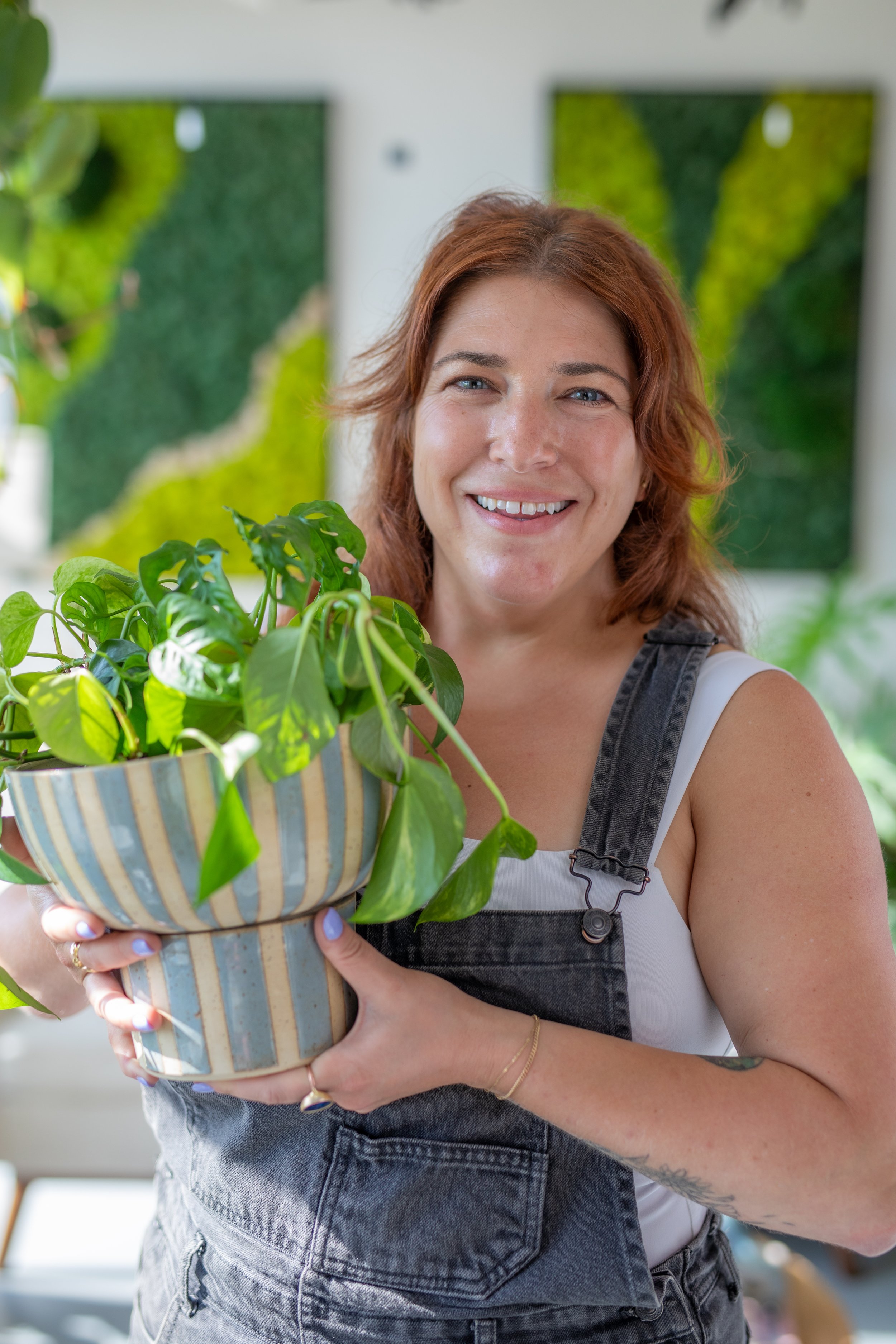 A smiling woman with red hair holding a potted plant with green leaves, standing indoors with a green wall art in the background.