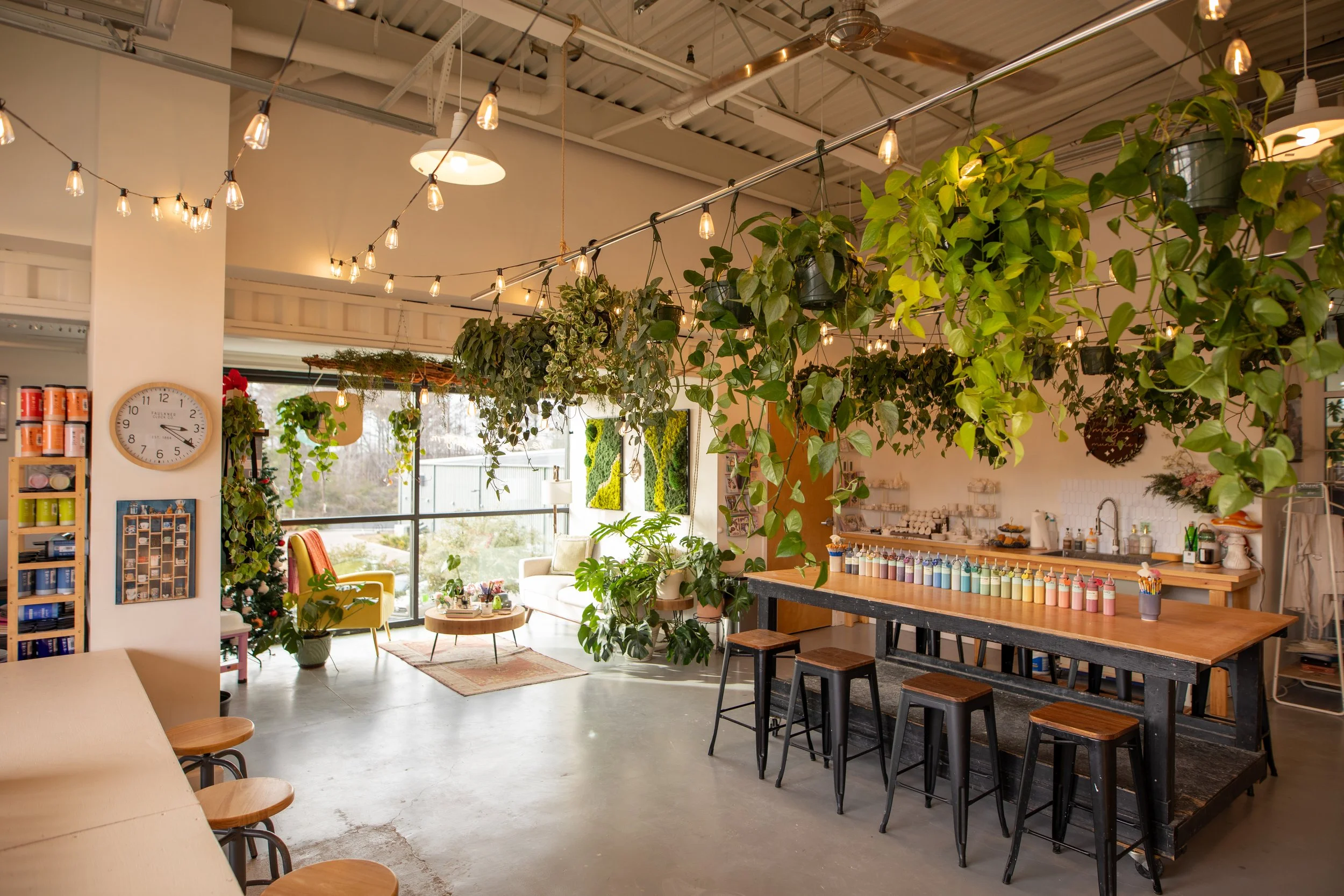 Interior of a cozy, well-lit store with hanging string lights, green hanging plants, a white ceiling, and a large window in the background showing outside trees. There are various displays including a round table with small plants, a colorful chair, and shelves with products. A wall clock is visible on a white pillar.