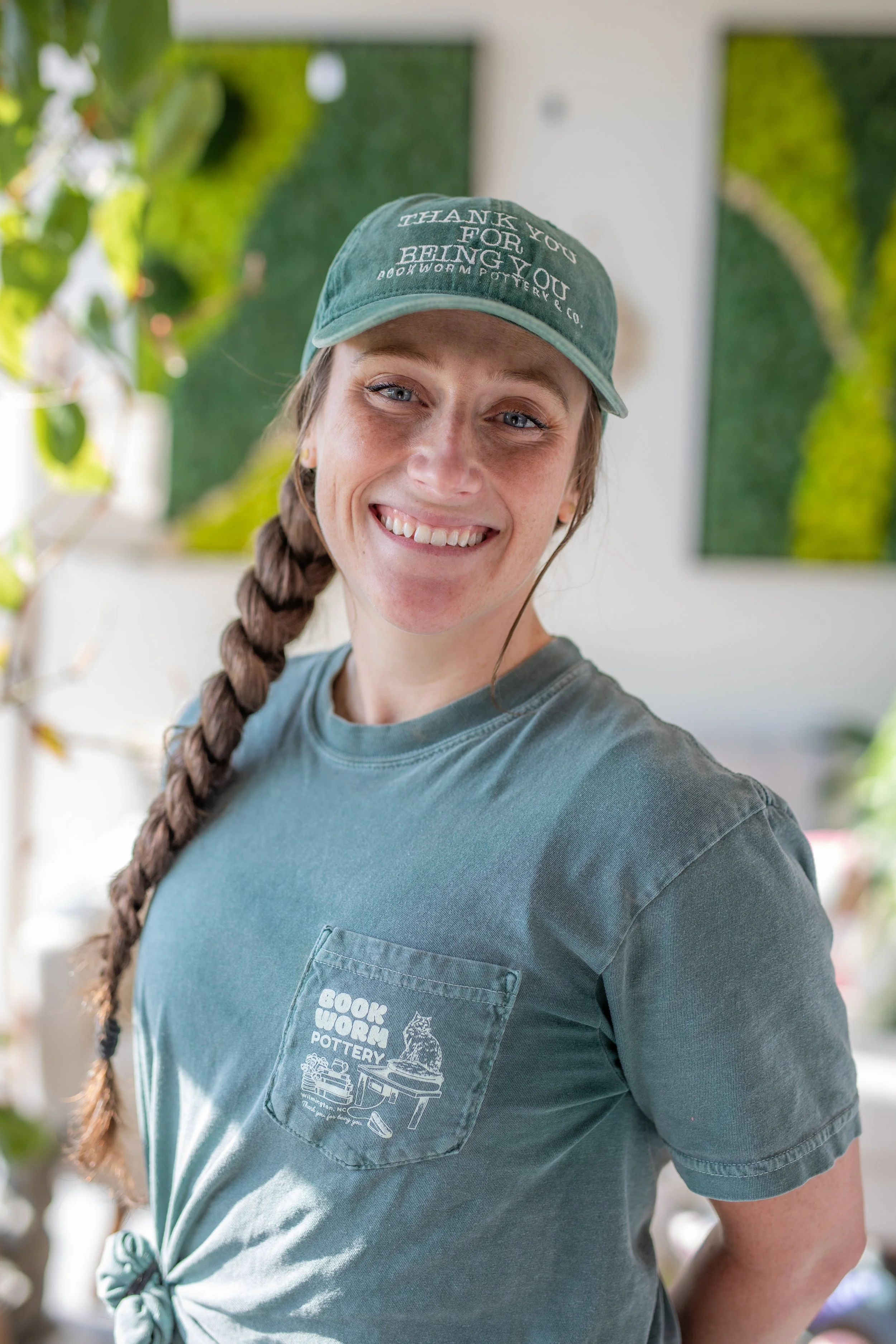 A smiling woman wearing a green baseball cap and a gray T-shirt with a pocket. The cap has text that says 'Thank you for being you' and a logo' The woman has light complexion, blue eyes, and a long braid over her shoulder. She is standing indoors with greenery and a white wall or art piece in the background.