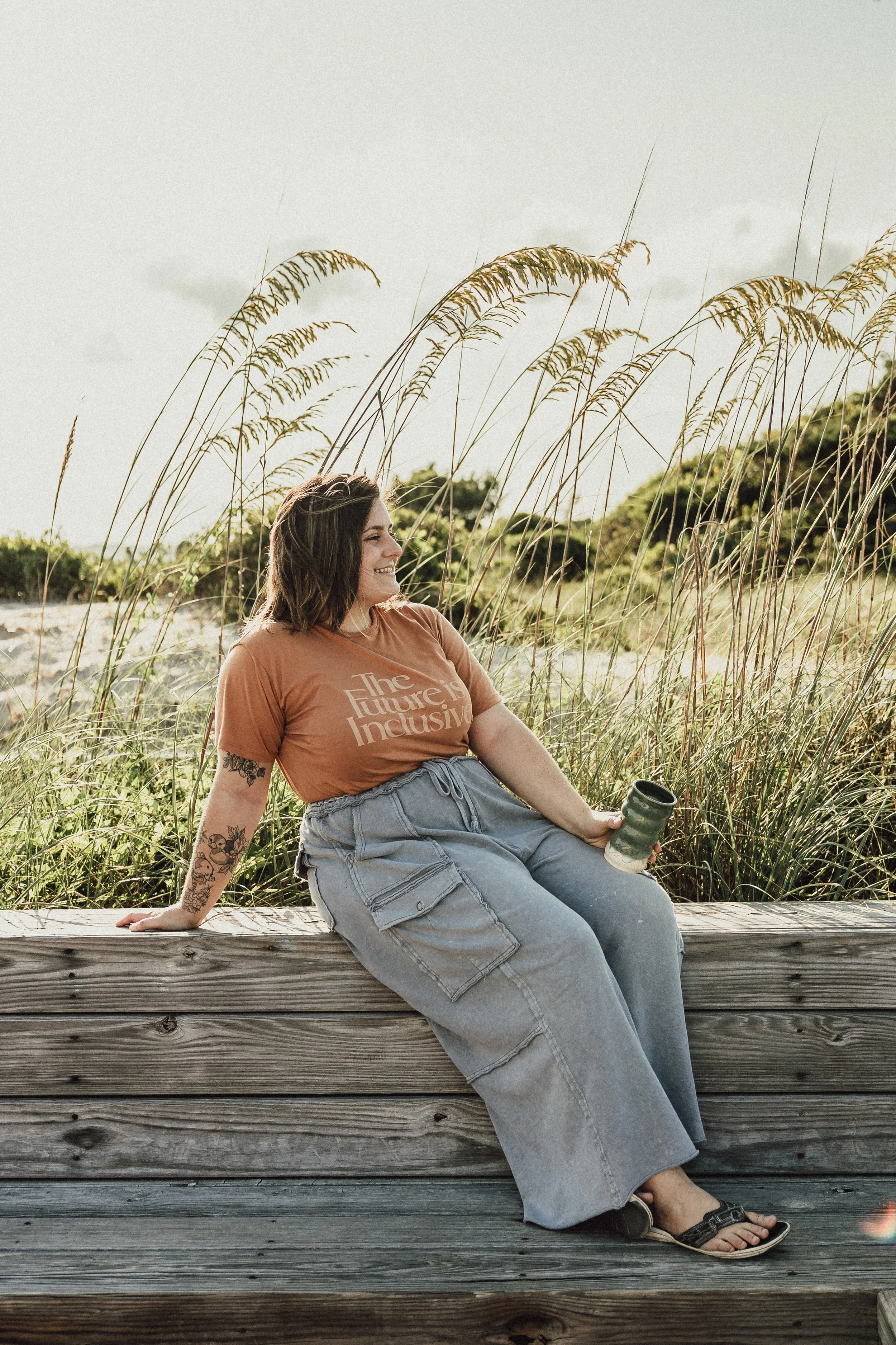 A woman with tattoos on her left arm sitting on a wooden bench outdoors during daytime. She is wearing a brown T-shirt that reads "The future is inclusive," light gray cargo pants, and black sandals. She is holding a green cup in her right hand and looking to her left with a smile. Tall grass and shrubs are in the background under a partly cloudy sky.