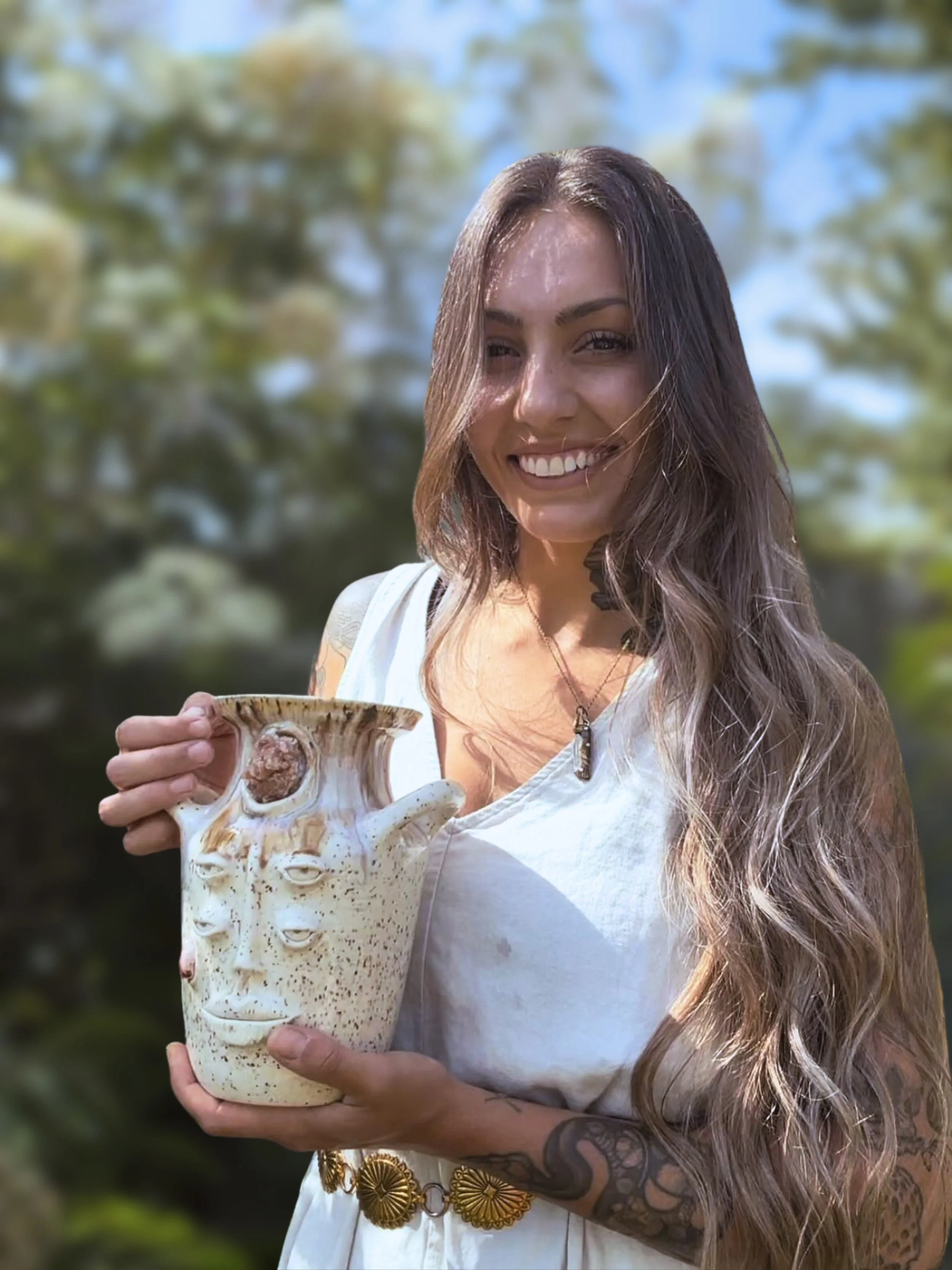 A woman with long wavy hair and tattoos on her arm holding a ceramic vase with a textured surface outdoors, with trees and a blue sky in the background.