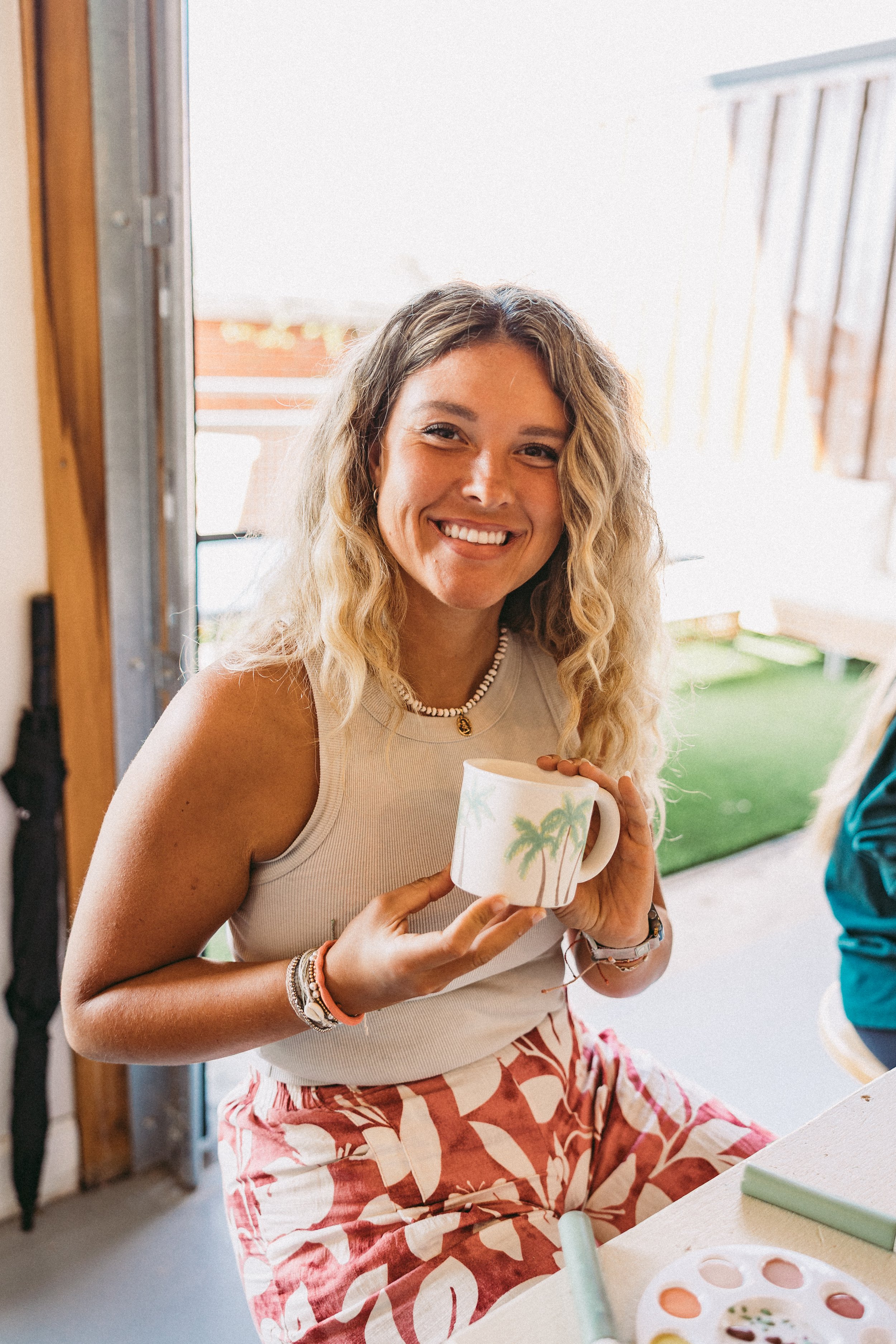 A woman smiling and holding a mug with a tropical design, sitting at a table inside a room with a door open to the patio.