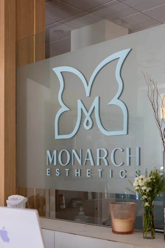 Sign for Monarch Esthetics with a butterfly logo on a glass wall, with flowers and a container on a counter in front.