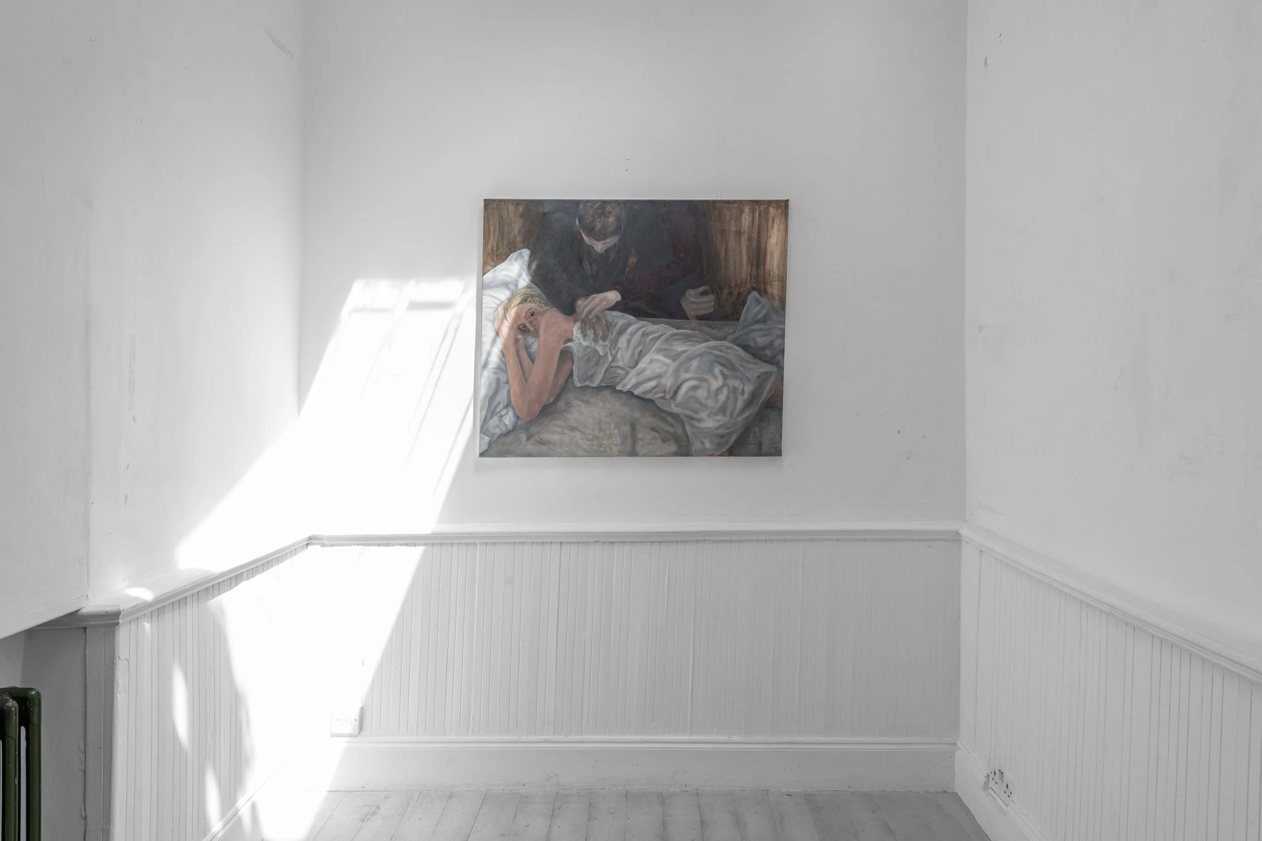 A painting of two people lying in bed, with one reading a book and the other looking on, mounted on a white wall in a bright room.
