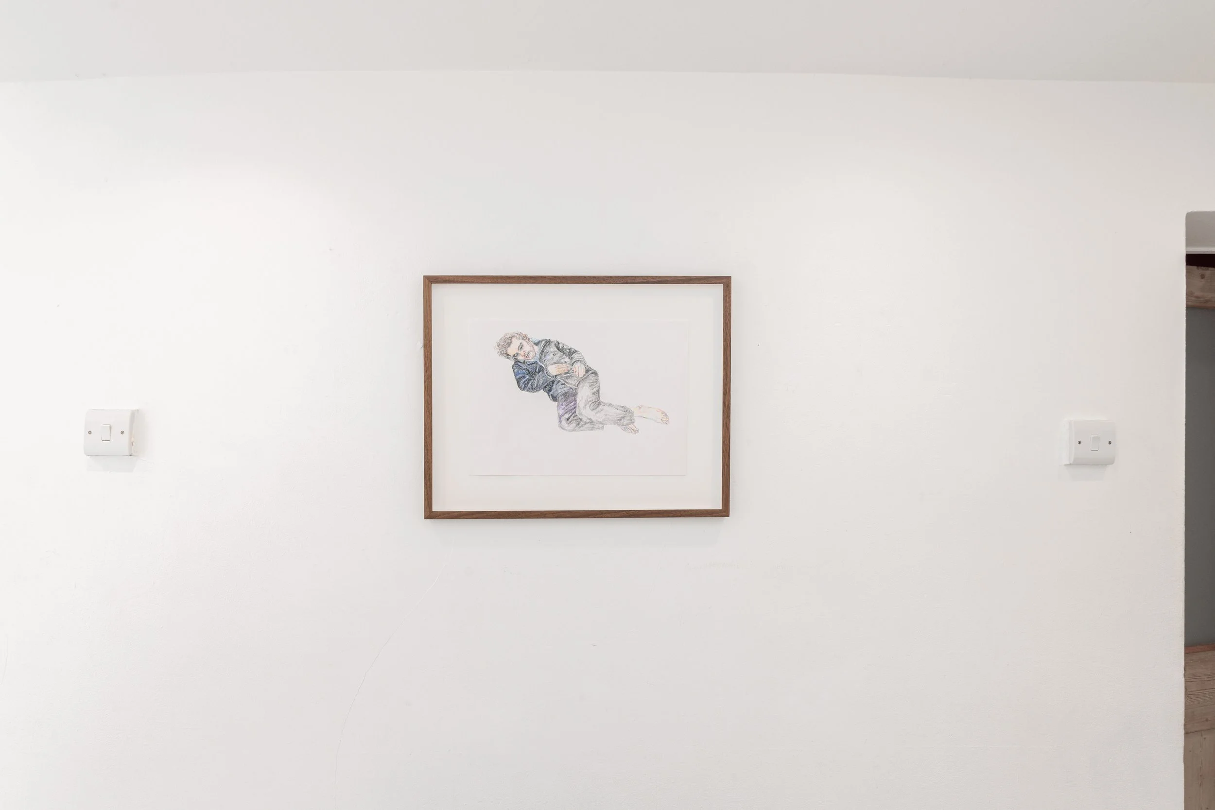 A framed drawing of a person with curly hair, wearing a dark jacket, sitting with arms crossed on their knee, on a white wall.