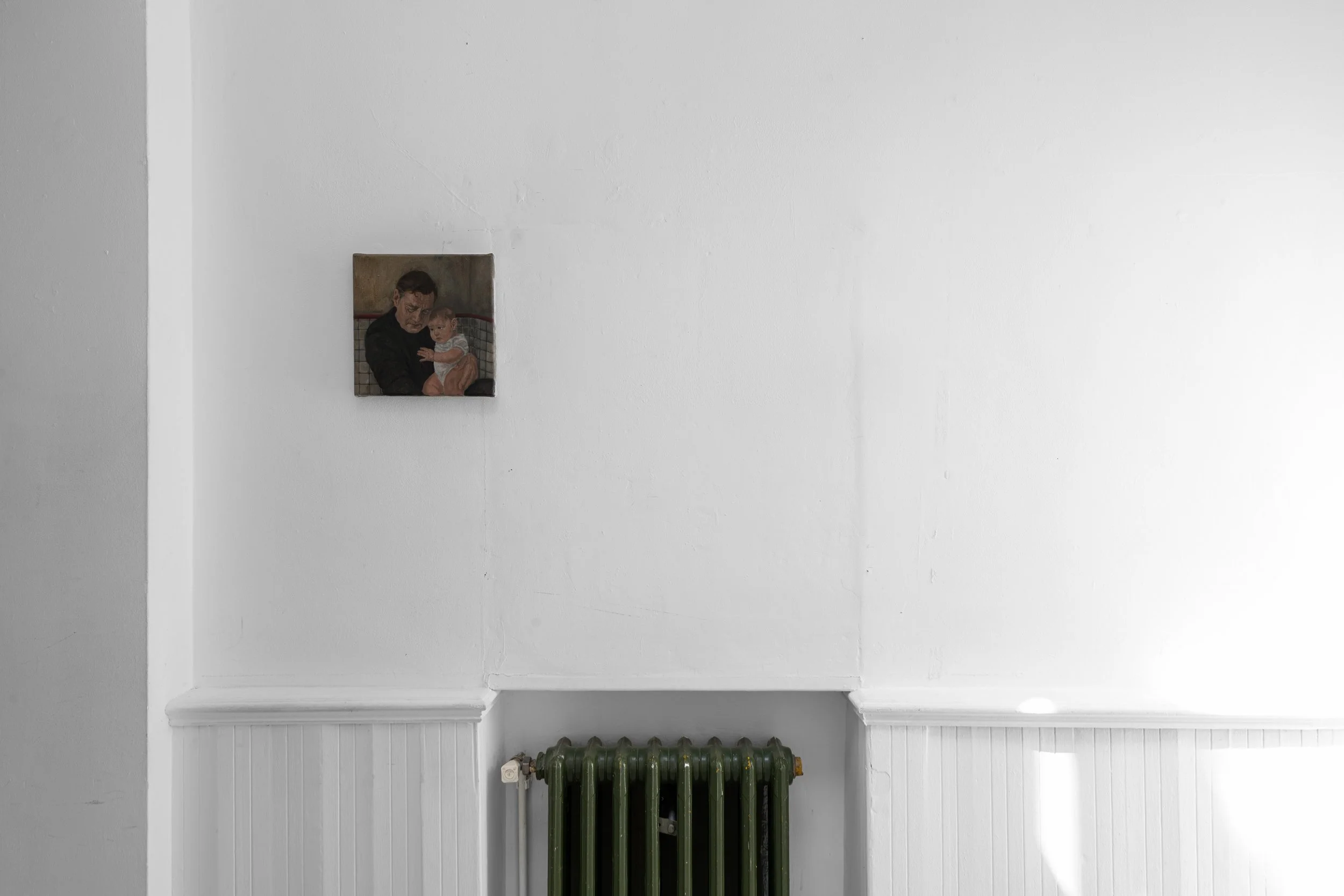 A small portrait painting of a man holding a baby is hung on a white wall. Below the painting is a green cast-iron radiator. The room has white wainscoting and a horizontal ledge at the bottom of the wall.