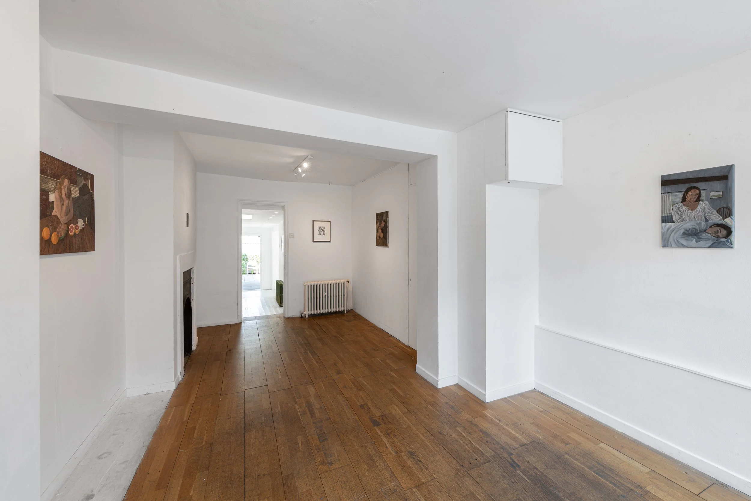 Interior view of a home with white walls and hardwood floors, featuring artwork hung on the walls, a radiator, and an open doorway leading to a brighter room outside.