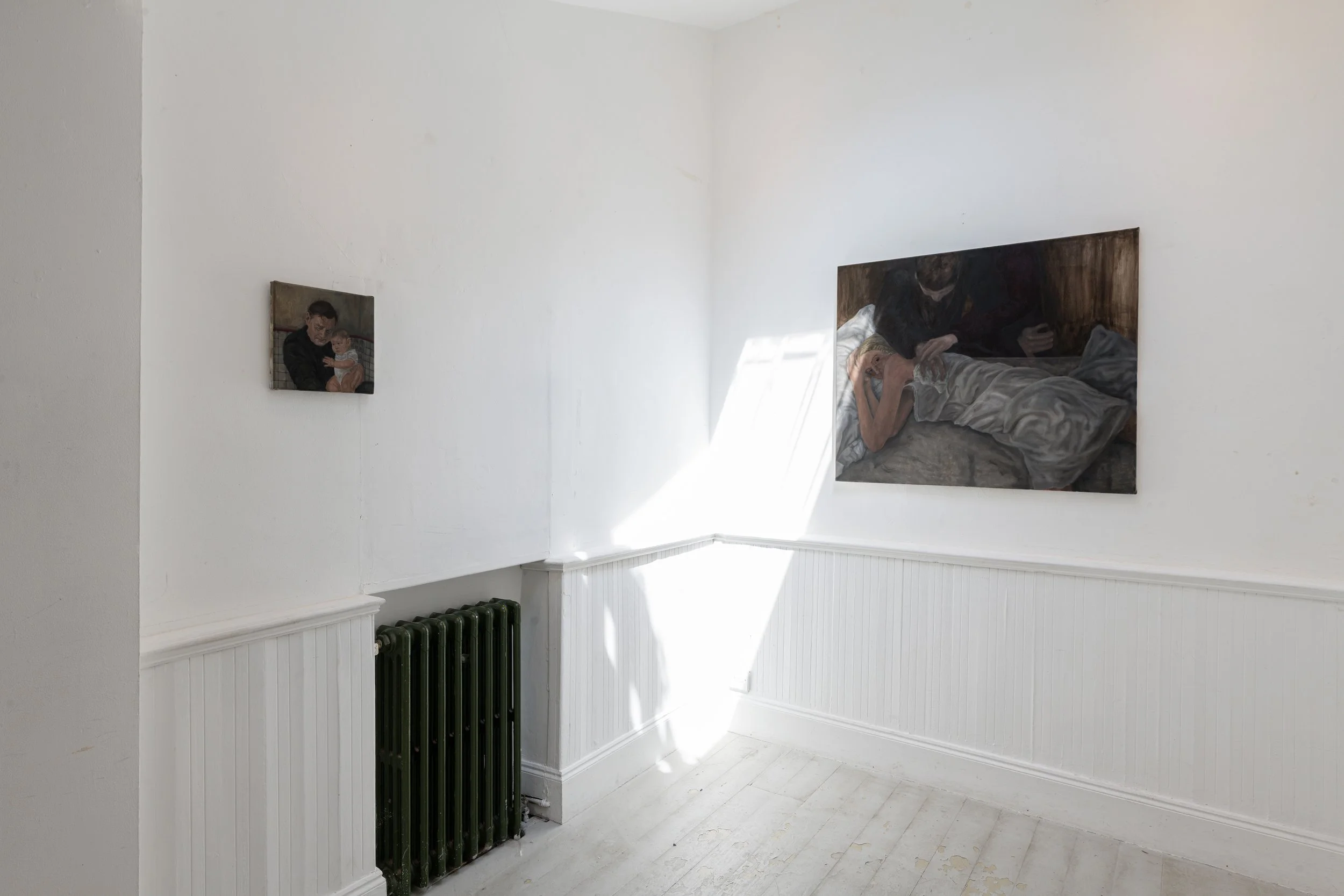 A white room with two paintings on the wall, one shows a man holding a baby, the other is a woman lying on a bed. There is a black radiator near the wall and a window casting sunlight on the wall.