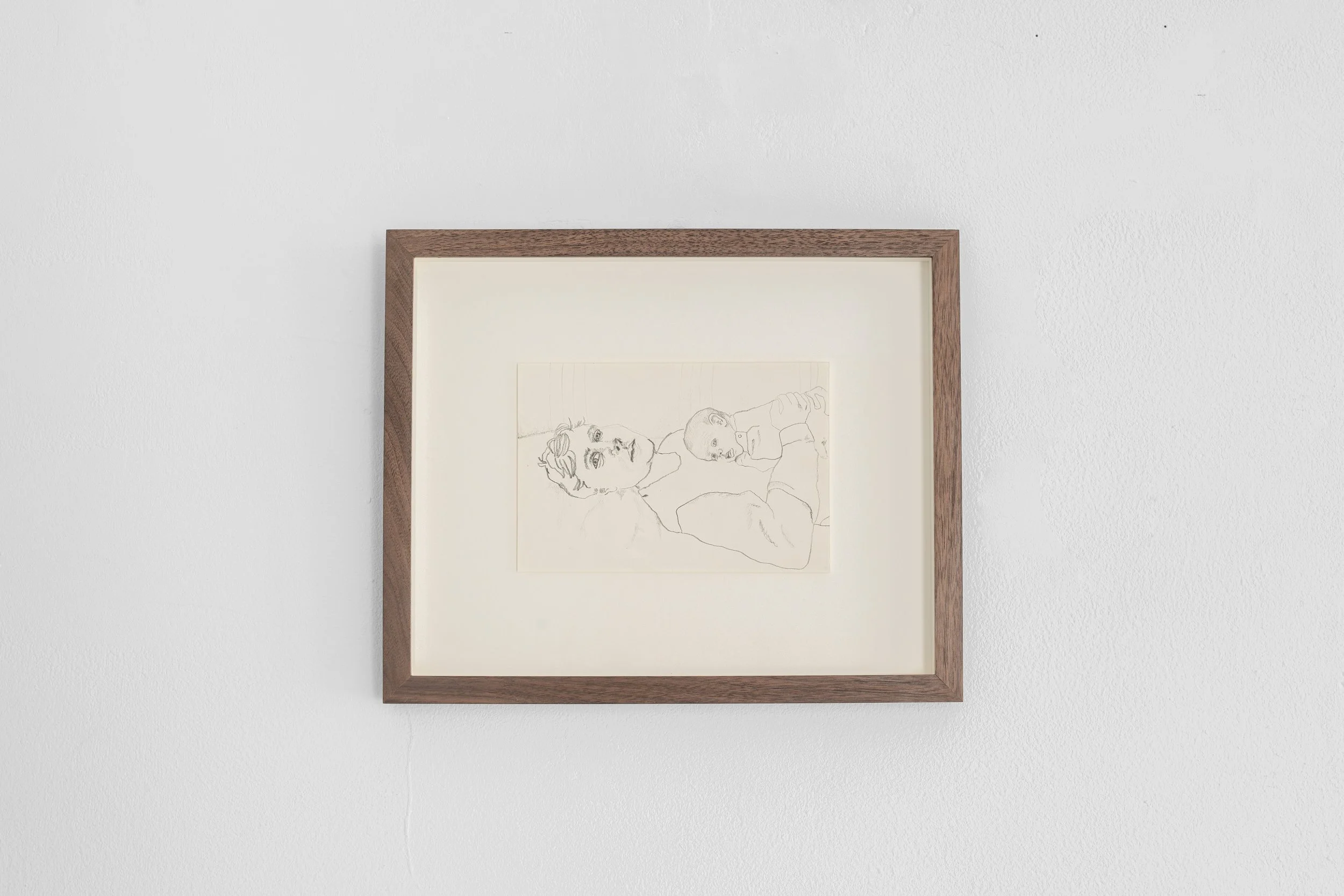 Framed sketch of two people lying down, one with short hair and a mustache, the other with a bald head, on a white wall.