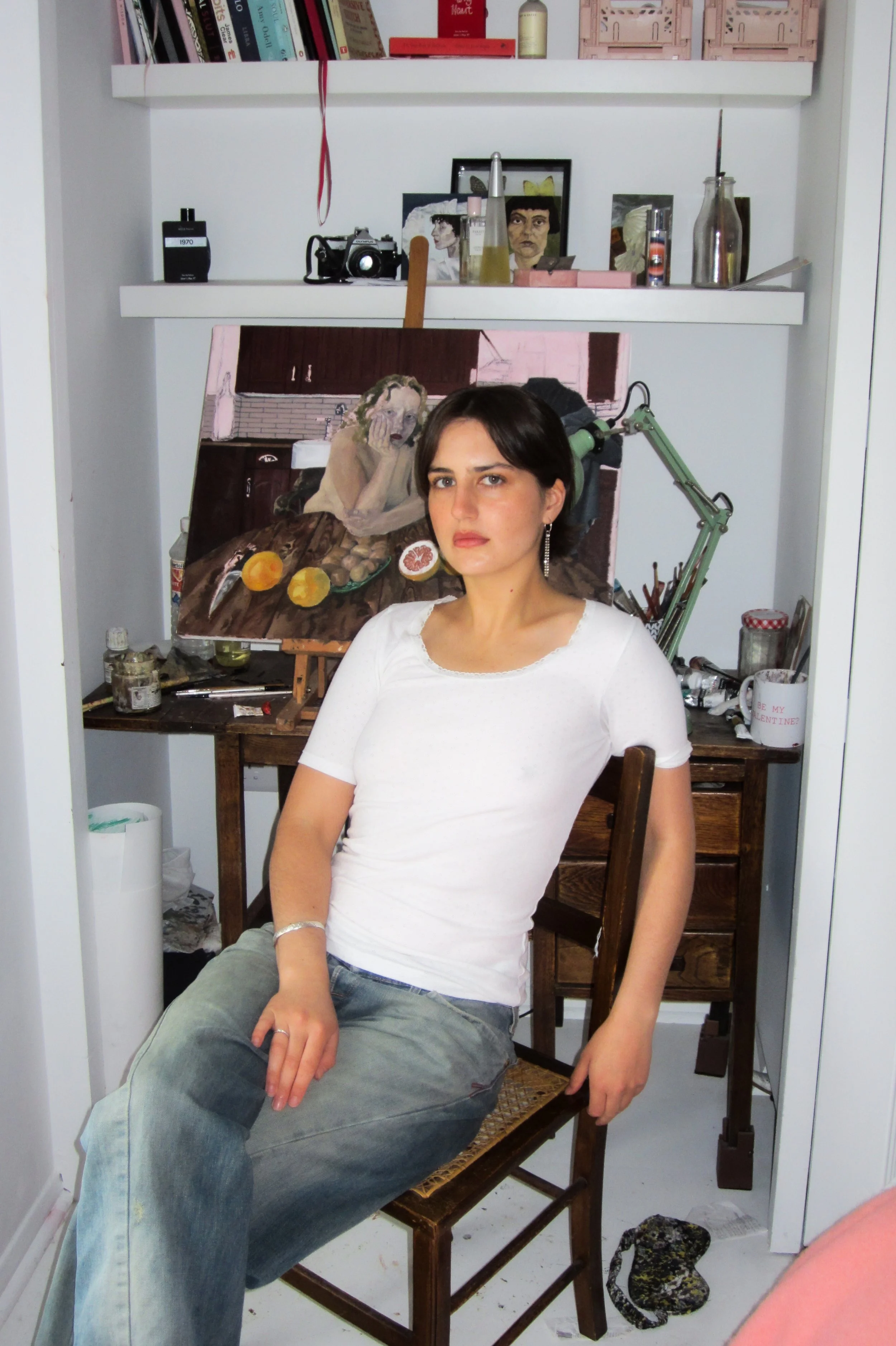 A woman with short dark hair, wearing a white shirt and gray pants, sitting on a wooden chair in art studio with her left arm resting on her knee. Behind her, a painting shows a woman with curly hair resting her face on her hand at a table with some food. The studio has various art supplies, a door, and a shelf with books and decorative objects.