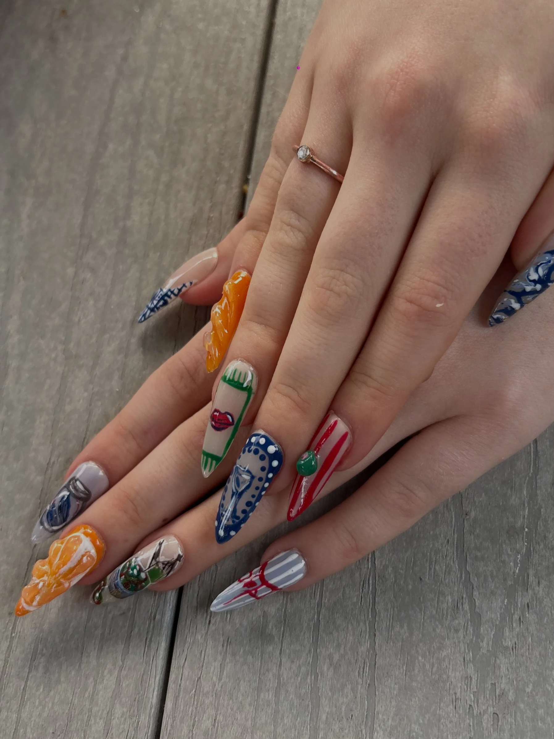 Close-up of a hand with long, decorated acrylic nails and a rose gold ring with a small gemstone.