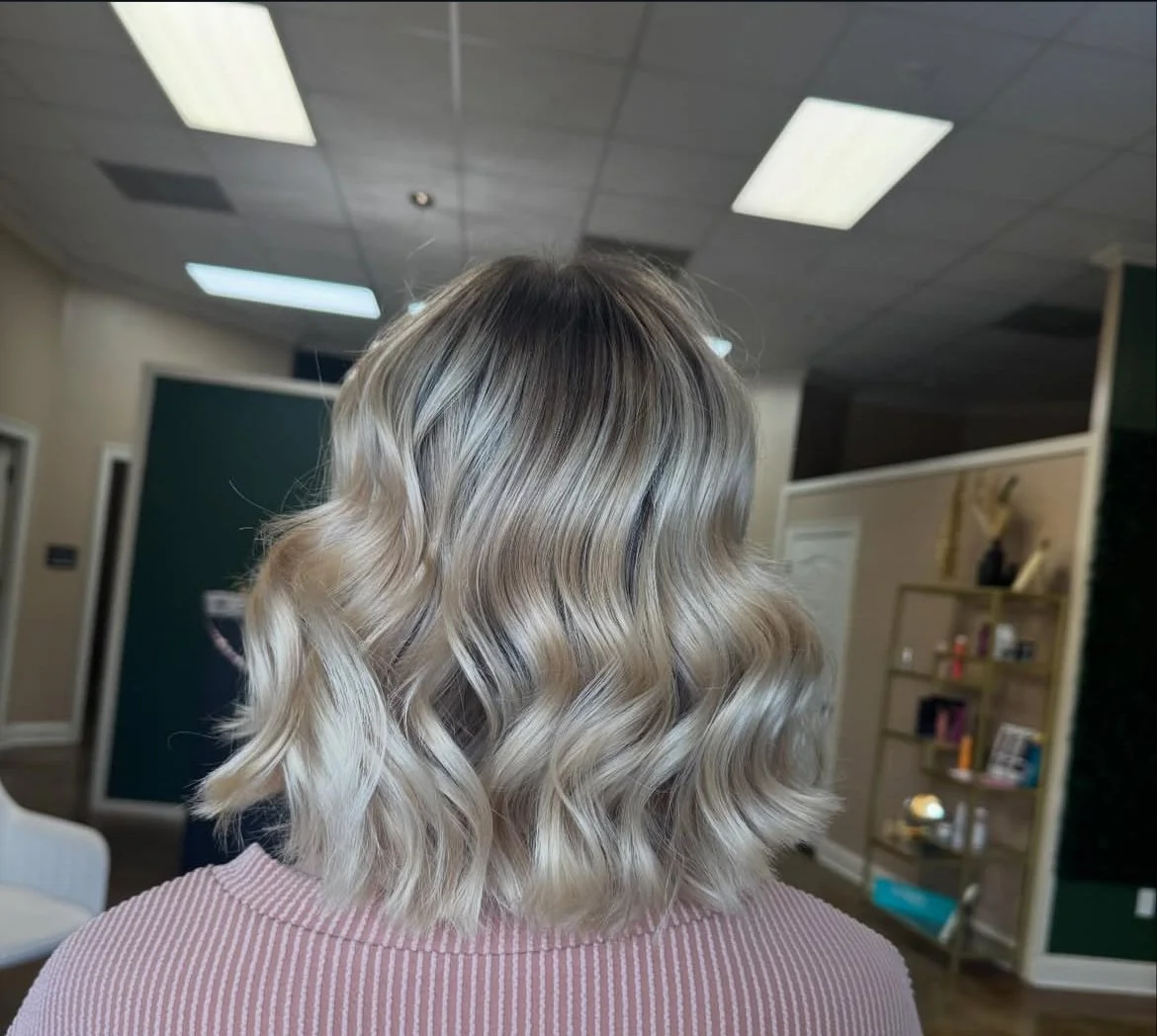 Back of a woman's head showing blonde, wavy shoulder-length hair in an indoor setting.