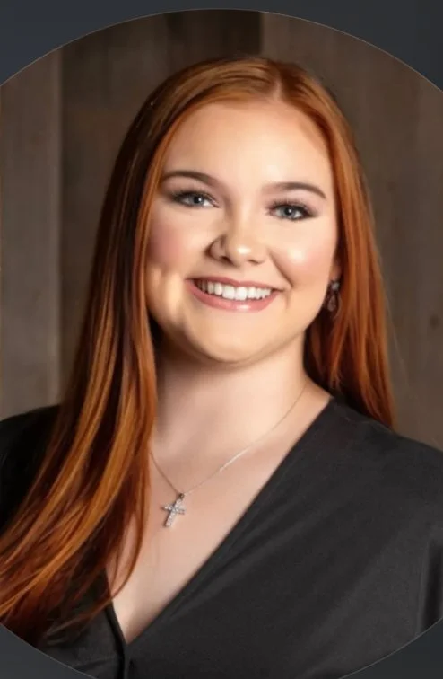 A young woman with long red hair smiling, wearing a black outfit, a necklace with a cross pendant, and earrings, standing against a wooden background.