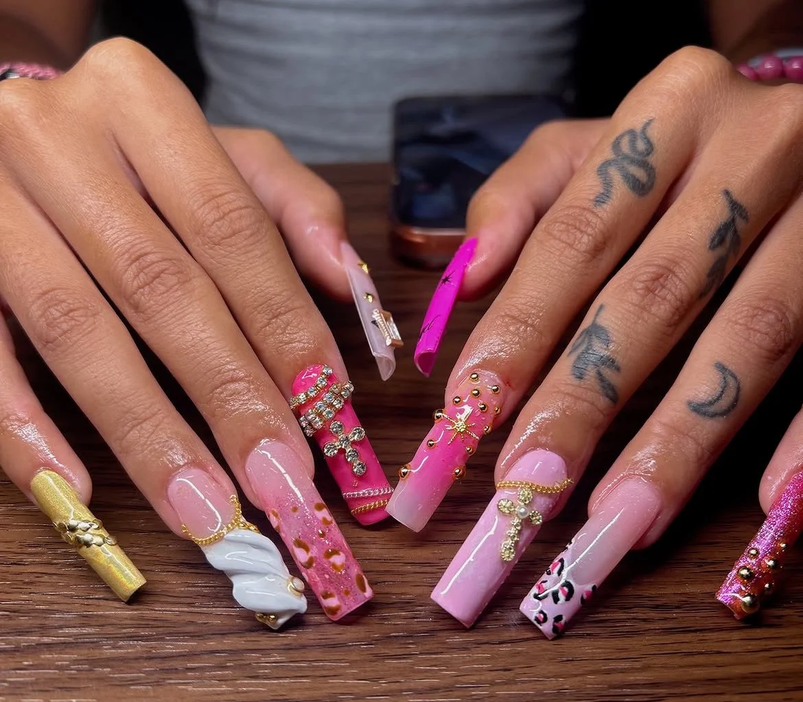 Close-up of hands with long, decorated acrylic nails featuring various designs, colors, and embellishments such as rhinestones and jewelry-style decorations, resting on a wooden surface.