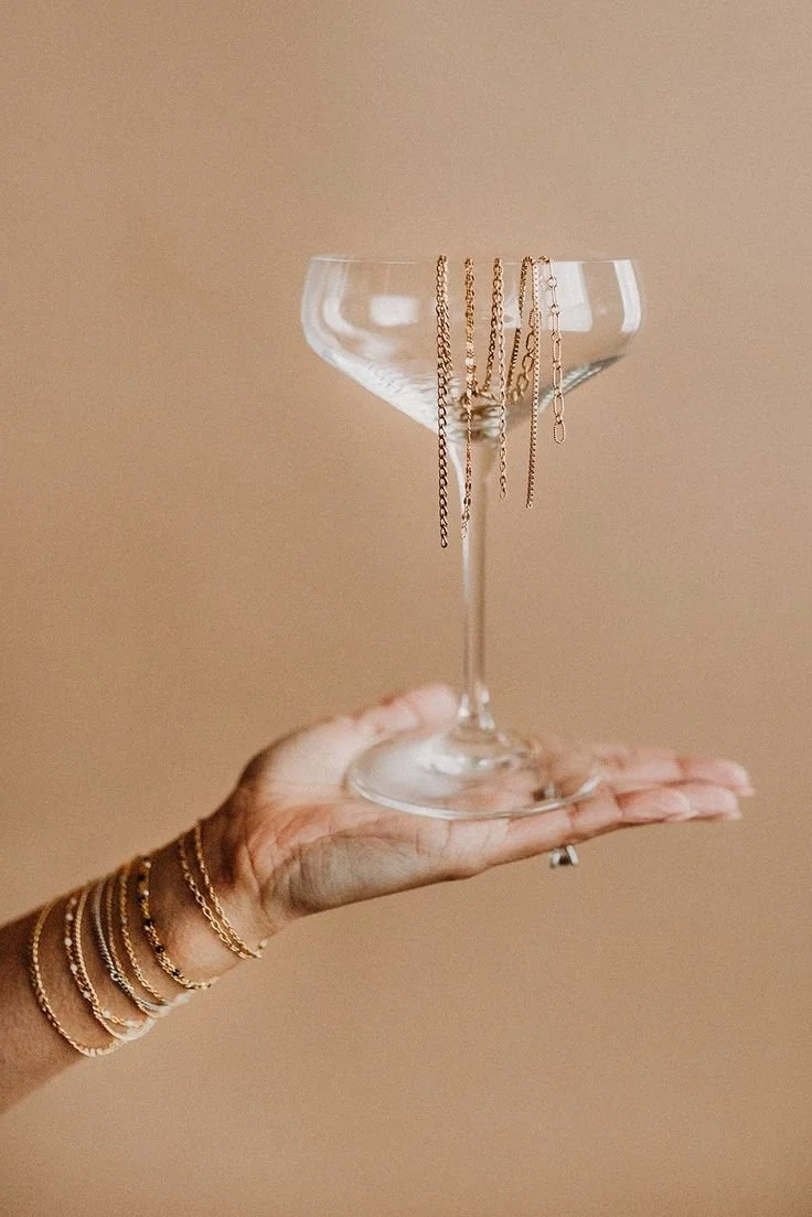 Hand holding a glass coupe with gold jewelry hanging on the rim and gold rings on wrist.