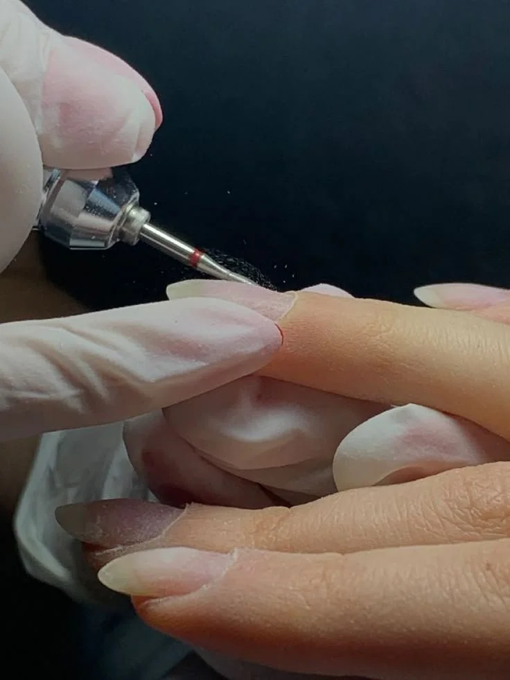 Close-up of a person receiving a manicure with a small electric drill on their fingernail, held by a nail technician wearing white gloves.