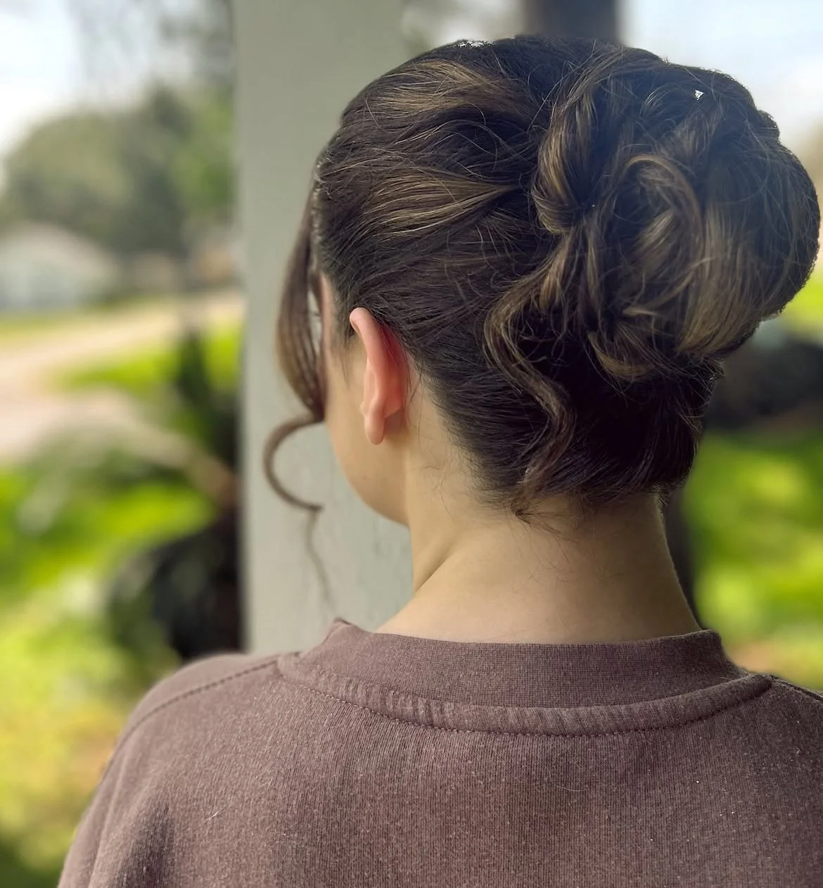 Rear view of a woman with a classic updo hairstyle with curls, wearing a brown top, outdoors with blurred greenery in the background.