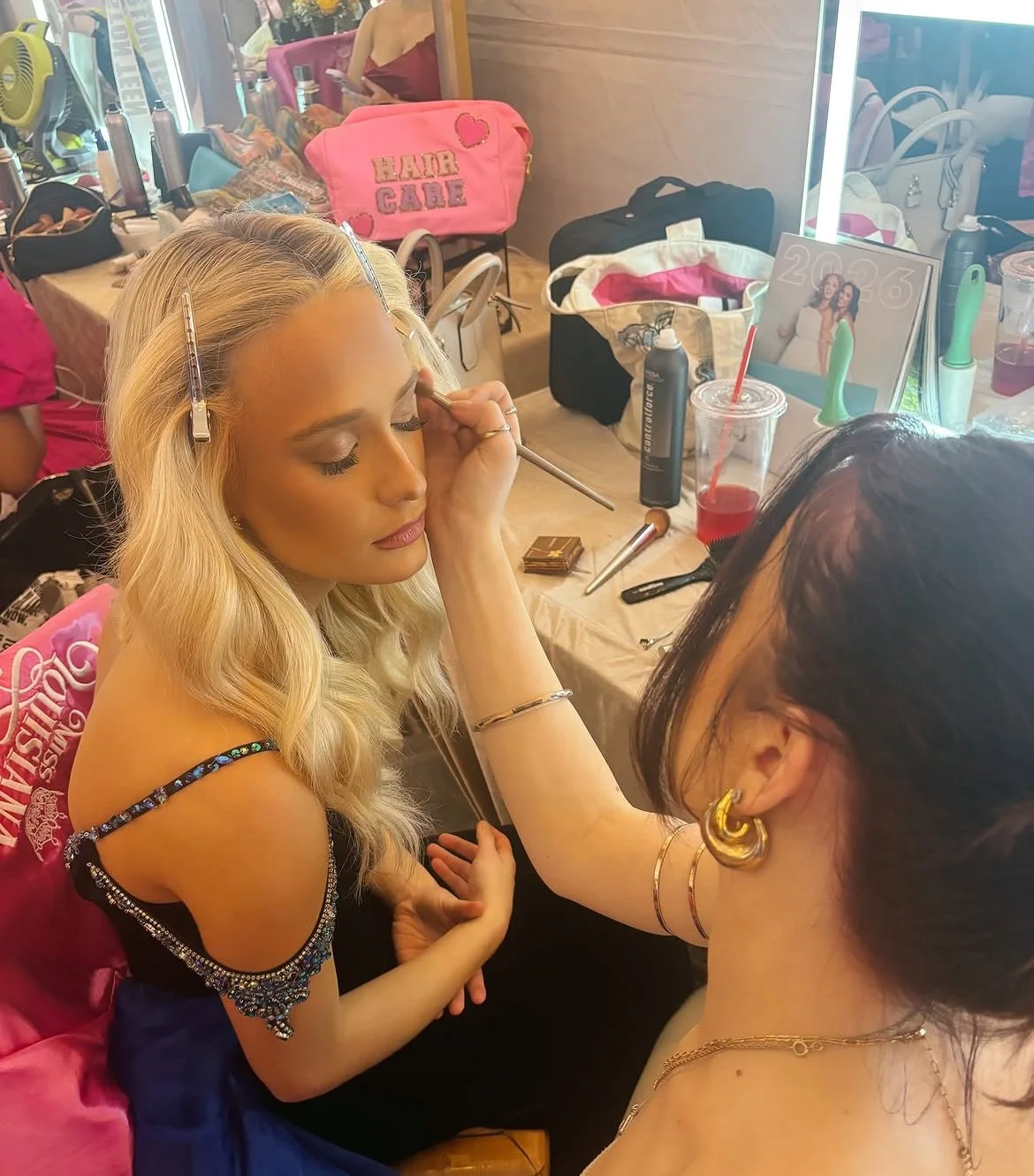 A woman with long blonde hair getting makeup applied by a makeup artist who is wearing gold jewelry and large gold earrings. The makeup artist is using a brush to apply makeup on the woman's face. The background shows makeup supplies, bags, a calendar, and drinks on the table.