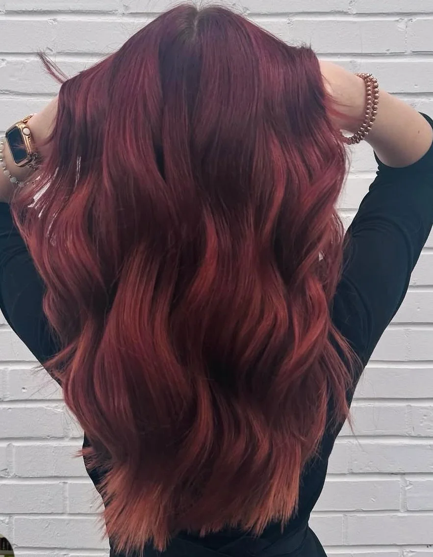 Person with long wavy red hair, wearing a black top, standing against white brick wall, with hands in hair.