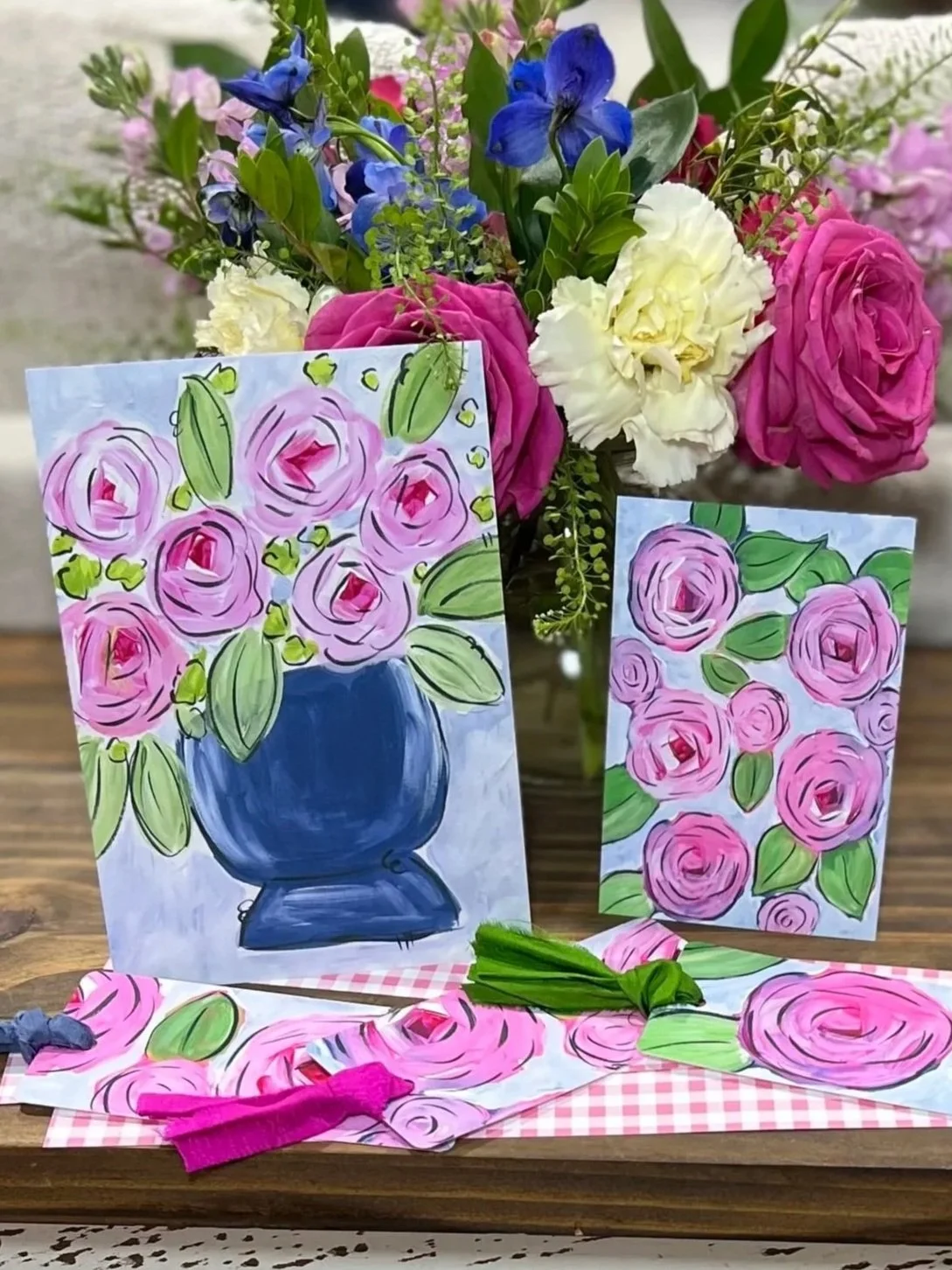A bouquet of real flowers with pink, white, and blue blooms, accompanied by two handmade art prints featuring pink rose illustrations, all displayed on a wooden table.
