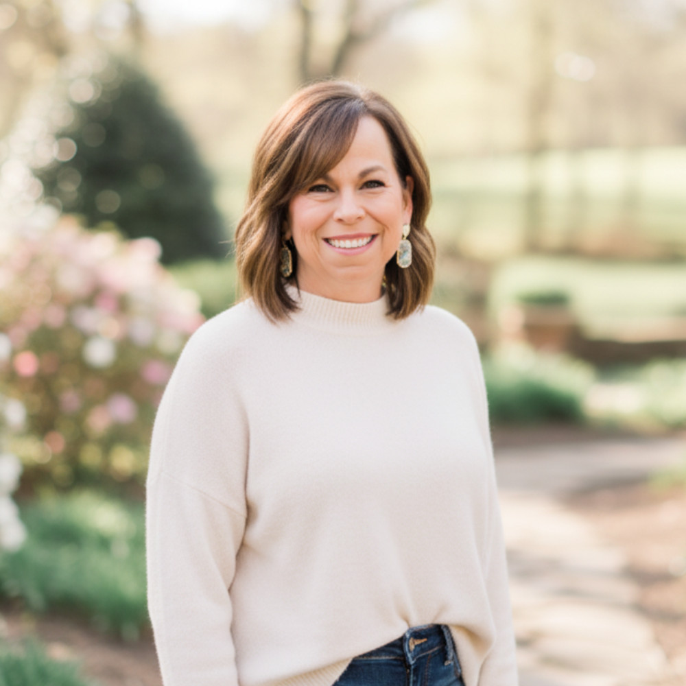 About page image - A woman with shoulder-length brown hair and earrings smiling outdoors in a garden or park setting.