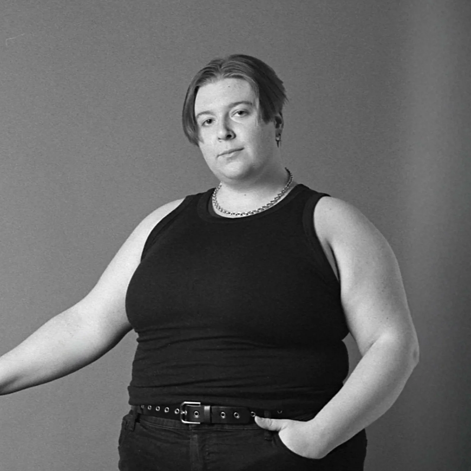 Black and white photo of a person with short, side-swept hair, wearing a sleeveless black top, a chain necklace, and earrings, against a plain background.