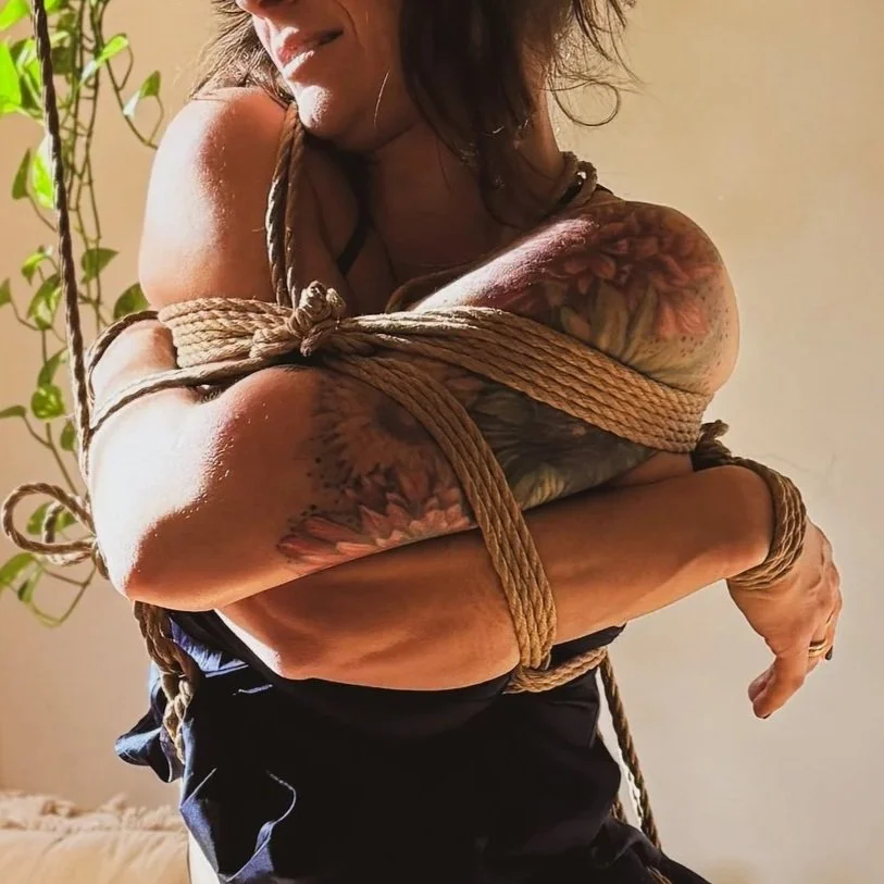 A woman with tattoos is bound with rope in a bondage pose.