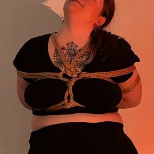 Woman with tattoos and dark clothing tied with rope, posing with her head tilted back.