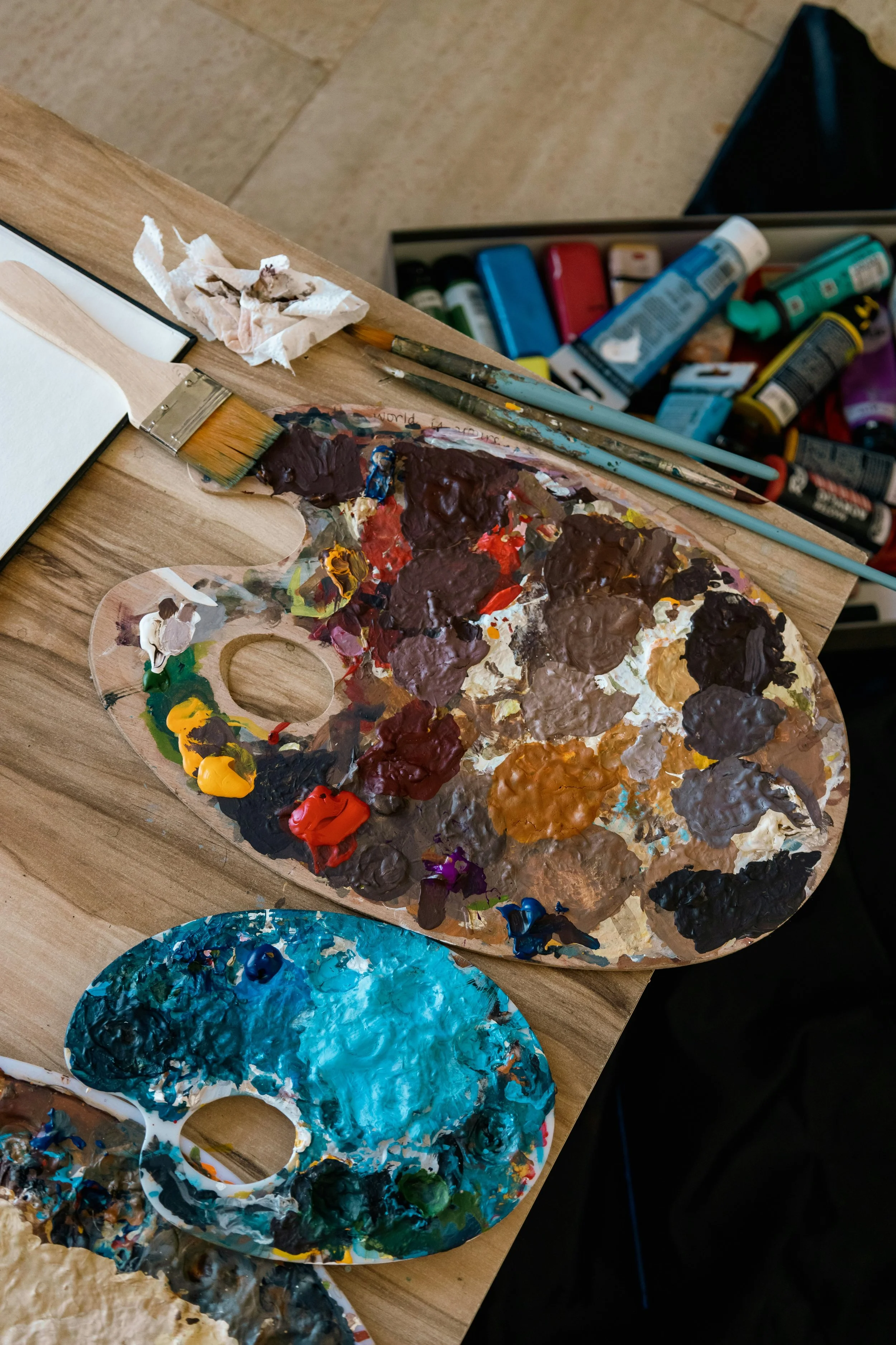 A mixing palette filled with various shades of blue, green, brown, and other colors of paint, placed on a wooden table. The scene also shows paintbrushes, a crumpled tissue, and a box of acrylic paint tubes in the background.