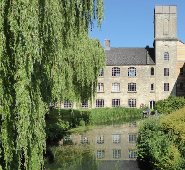 A historic stone building situated next to a calm river, with lush green trees and bushes surrounding it, and a bright blue sky overhead.