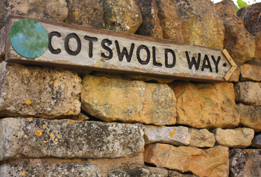 A wooden street sign reading 'Cotswold Way' mounted on a stone wall.