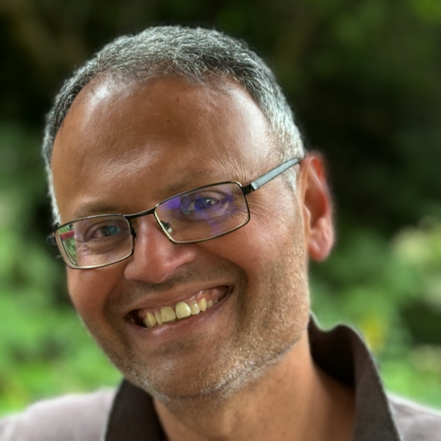 Close-up of a smiling man with glasses, short gray hair, and facial hair, outdoors with greenery in the background.