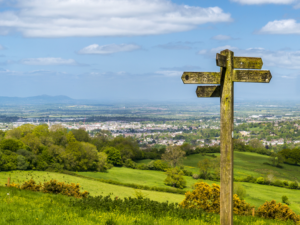 A wooden signpost with multiple directional signs is in a green hilly area overlooking a town with distant mountains in the background.
