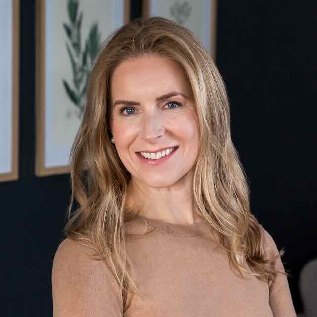 A smiling woman with long, wavy blonde hair, wearing a beige top, standing in front of framed botanical artwork on a dark wall.