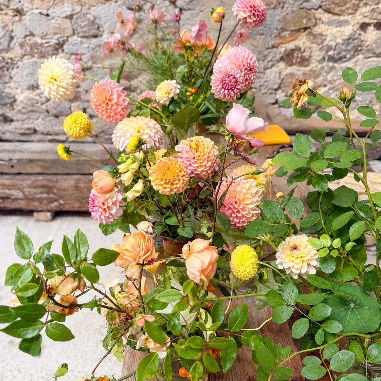 Pink, peach, yellow, and ivory flowers with loose greenery arranged in glass vessels along a wooden table against a stone wall.