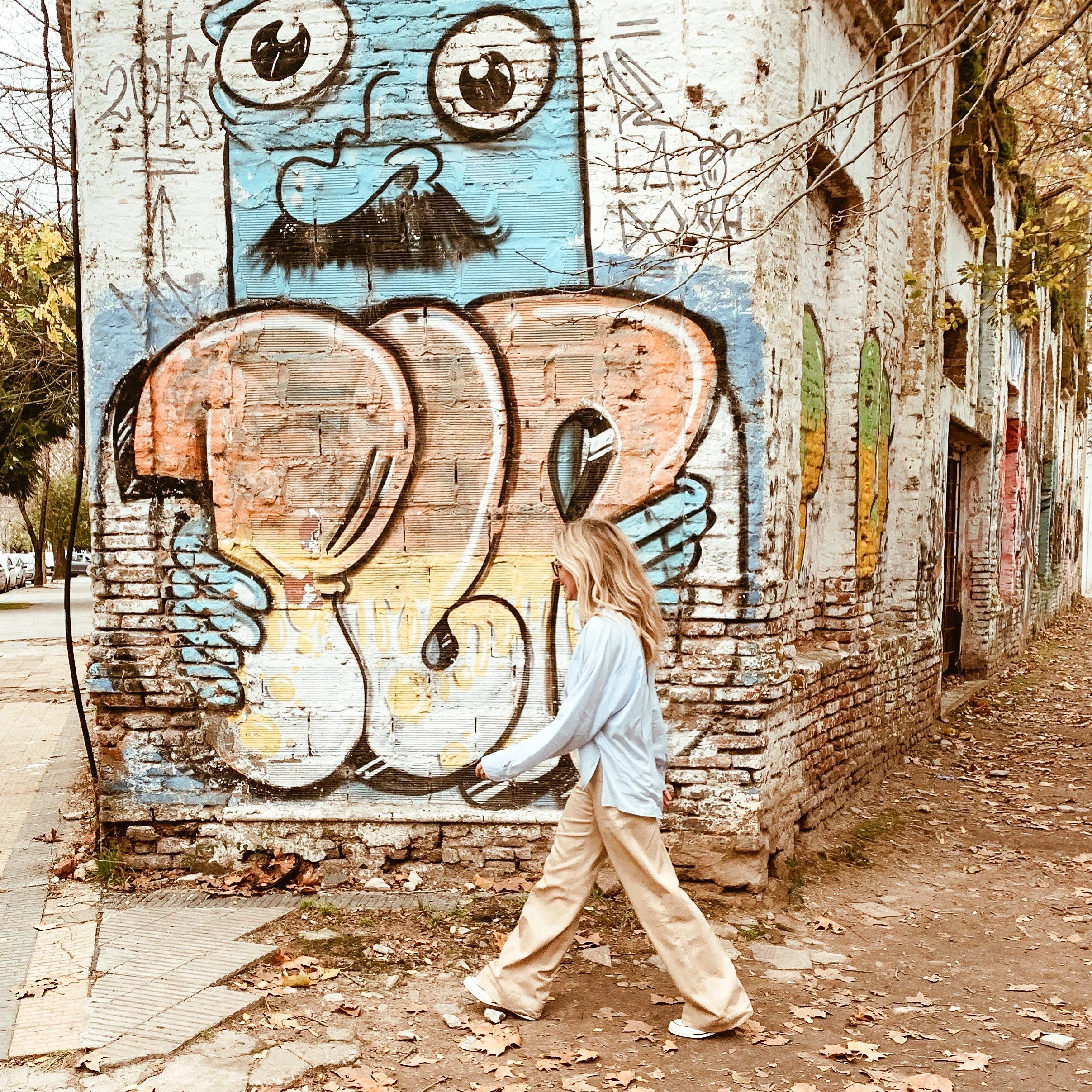 cMeg, founder of Meg&Co, walking past a colorful street-art mural in a light blue shirt and beige trousers.