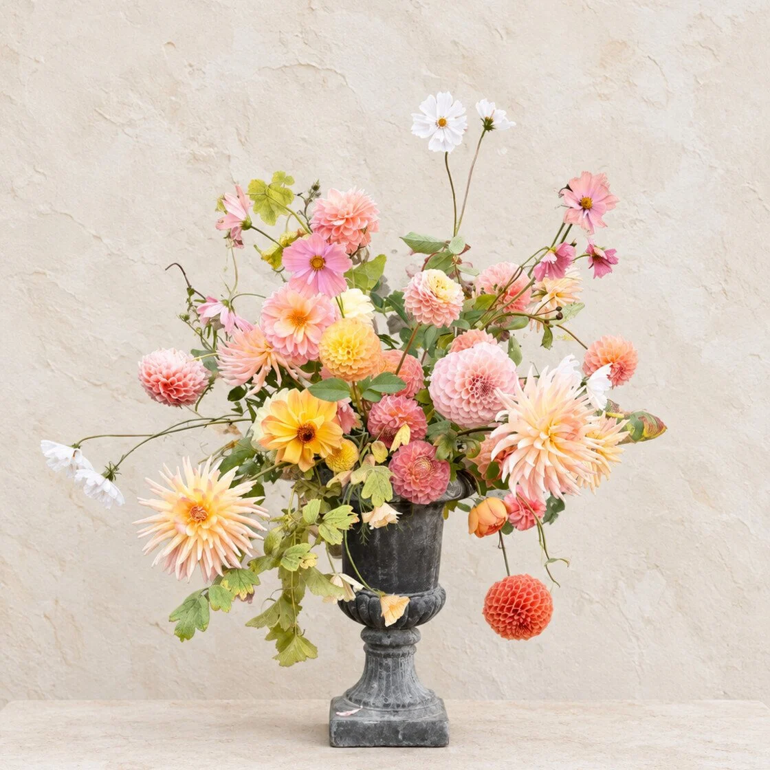 Loose pink, peach and yellow garden flowers arranged in a weathered black urn by Meg&Co