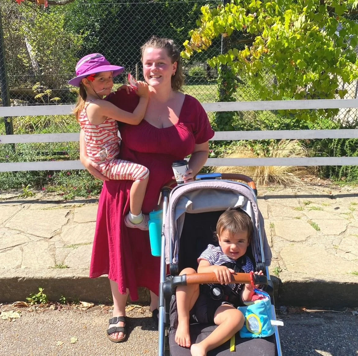 Lara Diprose, postpartum doula, holding her daughter, her son sitting in a buggy, outdoors on a sunny day in Folkestone, Kent.