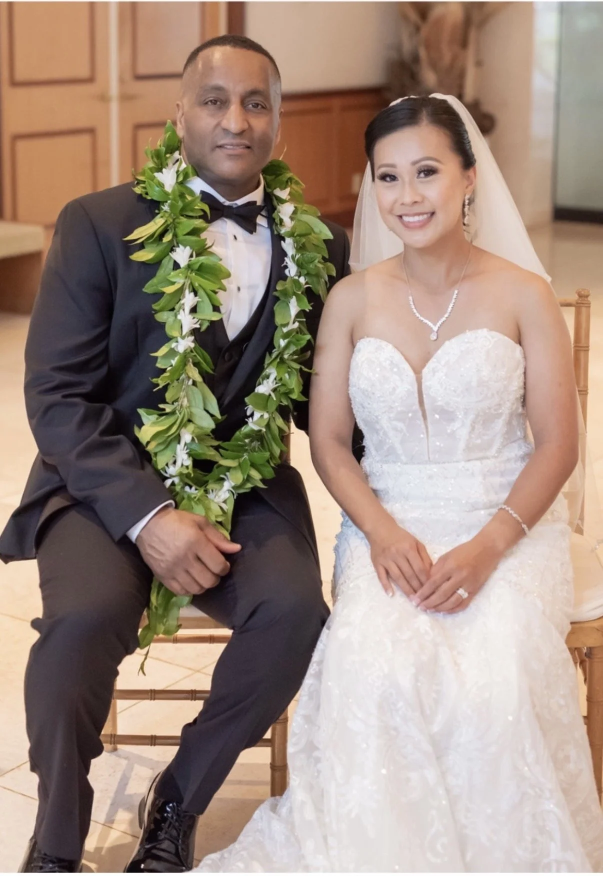 A bride and groom pose for a wedding photo inside a building. The groom wears a black tuxedo with a white shirt, black bow tie, and a green lei with white flowers. The bride wears a white strapless wedding gown with lace details, a pearl necklace, and a veil. They are sitting on wooden chairs, smiling at the camera.
