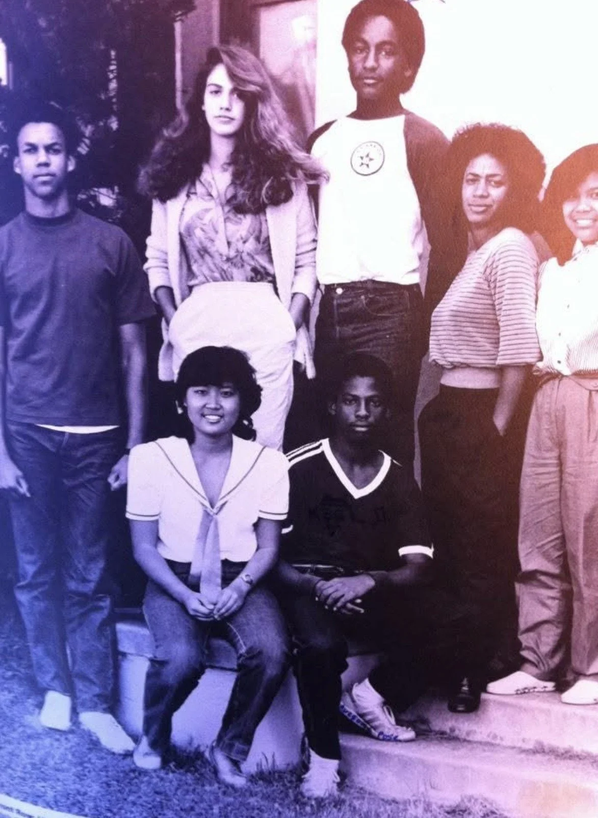 Group of seven diverse young people posing outdoors for a photo, some sitting, some standing, in casual clothing.