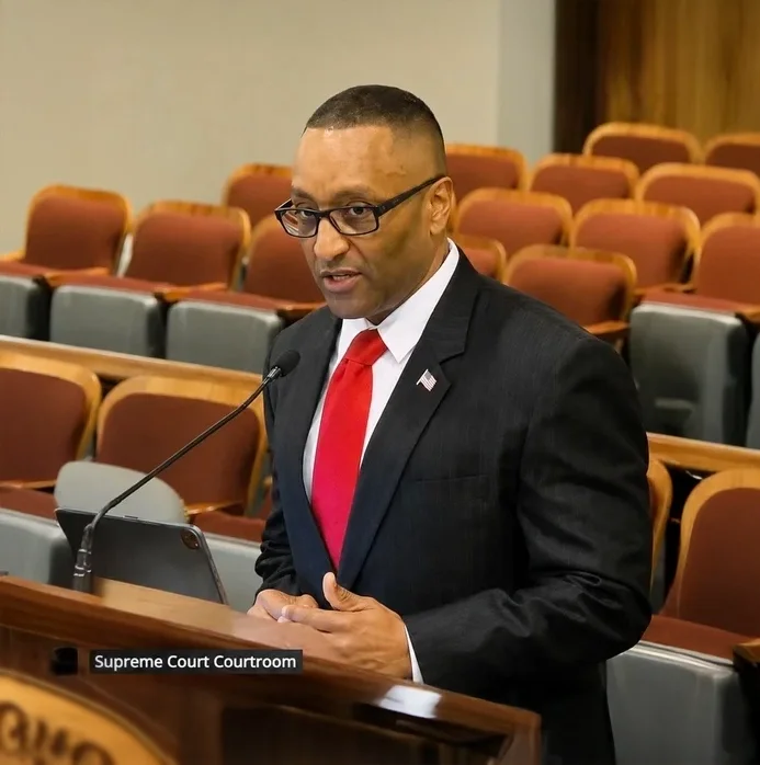 A man in a dark suit, white shirt, and red tie speaking at a podium in a Supreme Court courtroom. He is wearing glasses and standing behind a laptop, with empty seats in the background.