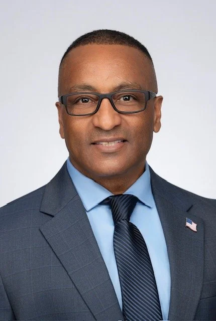 Portrait of a man wearing glasses, a blue suit, light blue shirt, and a dark tie with a small American flag pin on his lapel, against a light gray background.