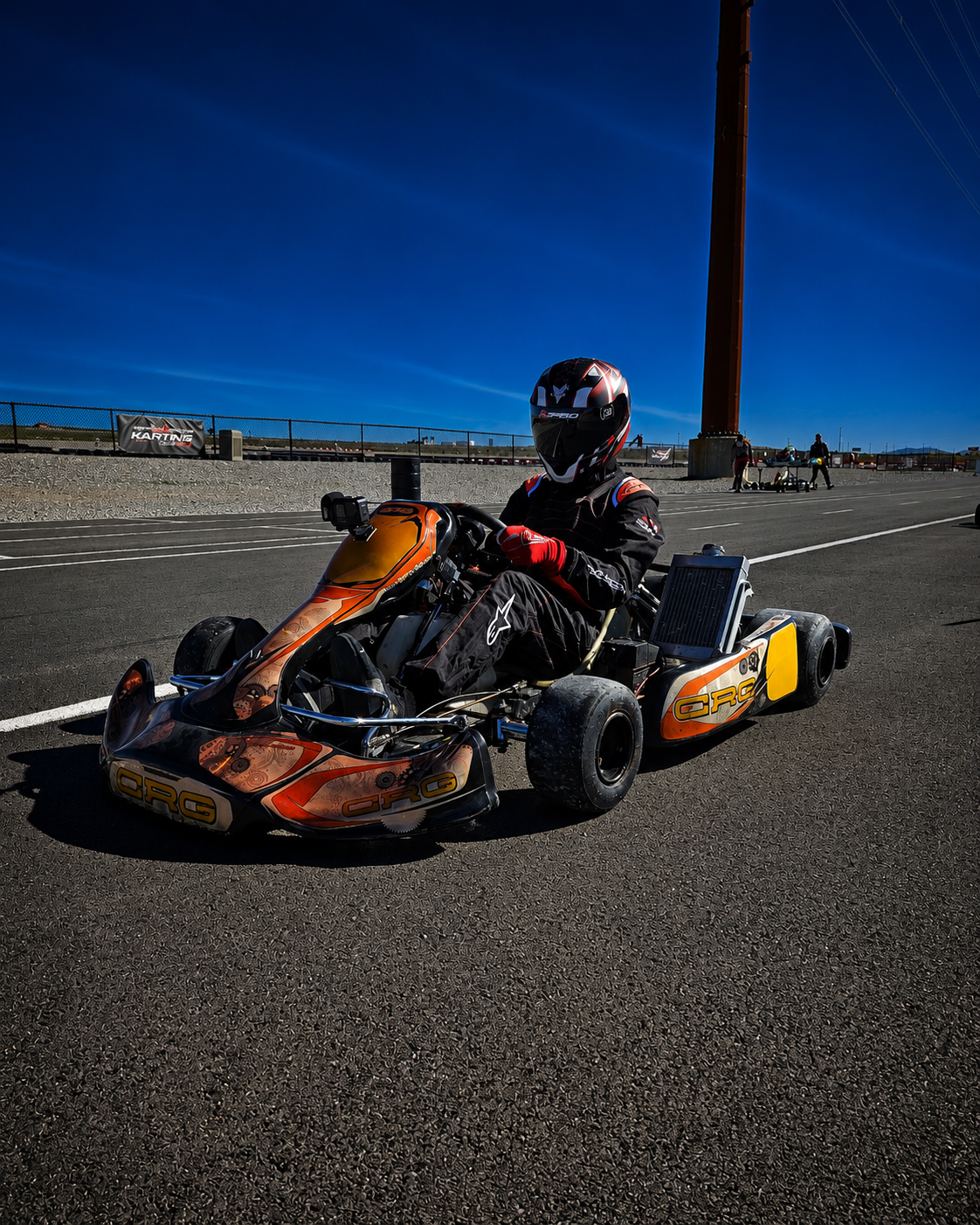 A person wearing a black racing suit and helmet sitting in a go-kart on a race track with a clear blue sky in the background.
