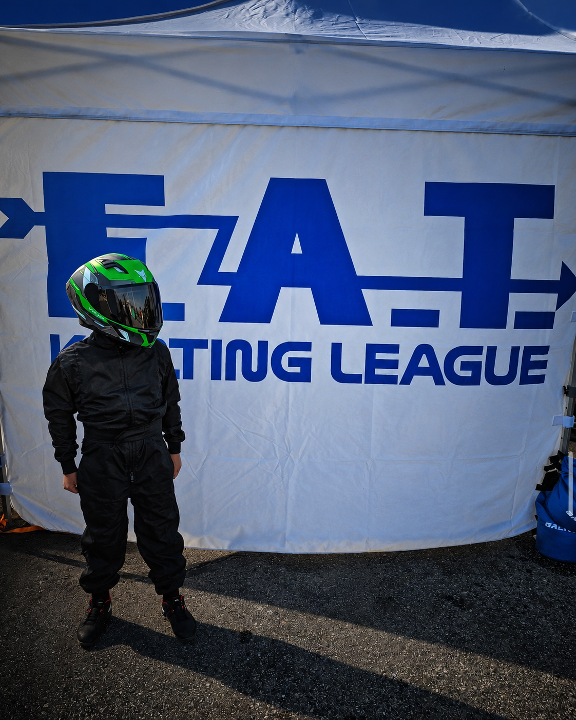 A person in a black racing suit and green helmet standing in front of a large white banner with blue text that reads 'EAT RACING LEAGUE'.