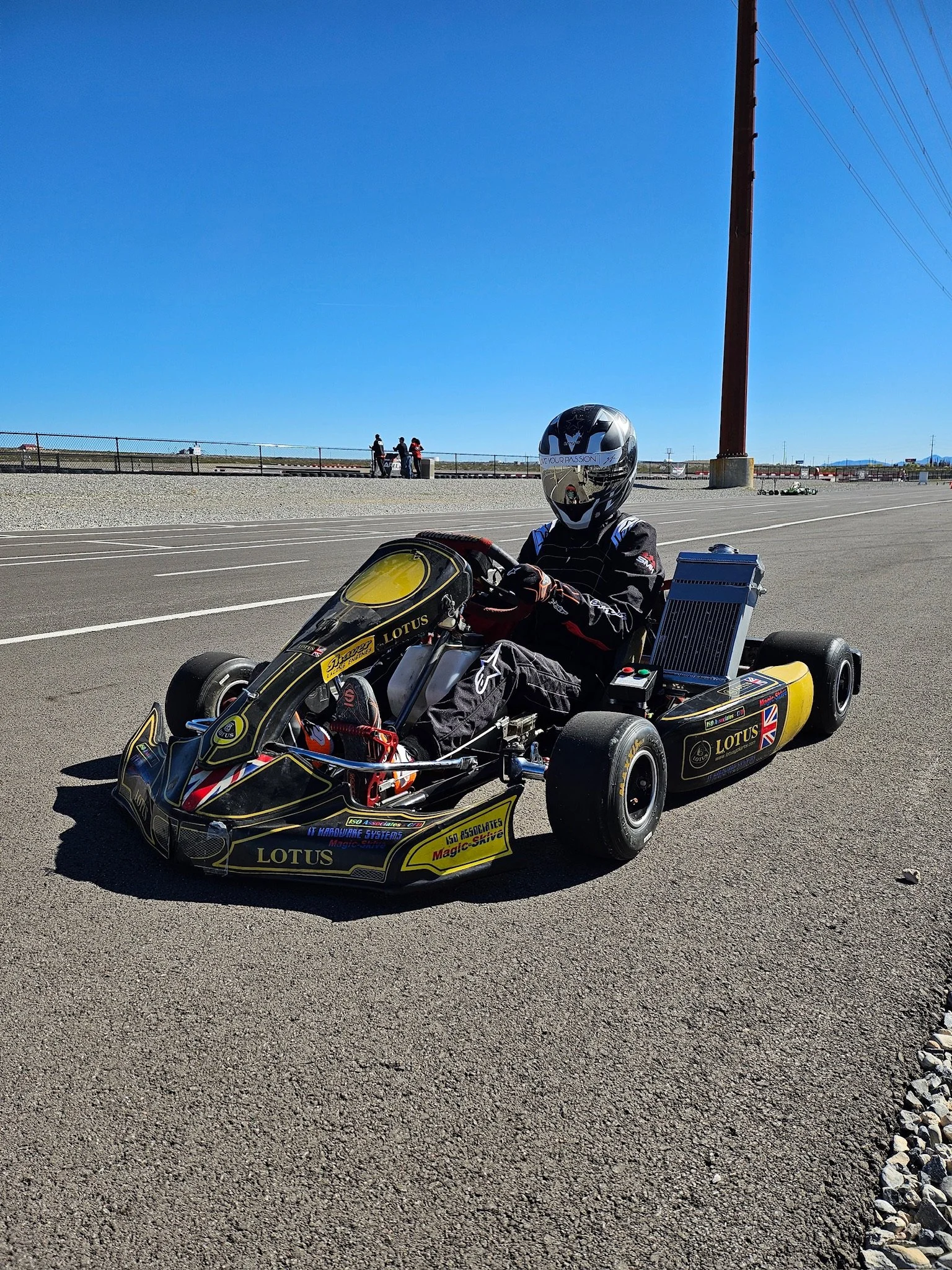 A person wearing a helmet and racing suit sitting in a go-kart on a paved track under clear blue sky, with a large red support beam and power lines in the background.
