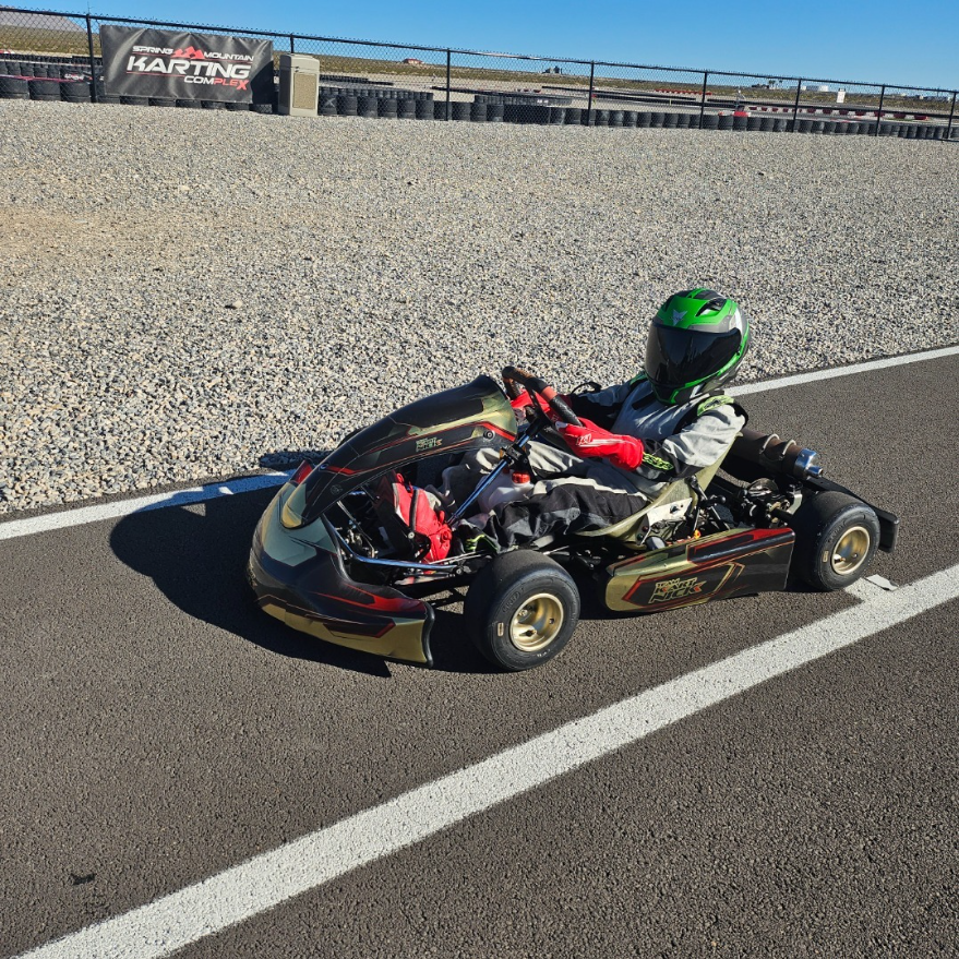 Child in a go-kart wearing a green helmet and red gloves, sitting on the starting line of a racing track with a banner that says 'Spring Mountain Karting Complex' in the background.