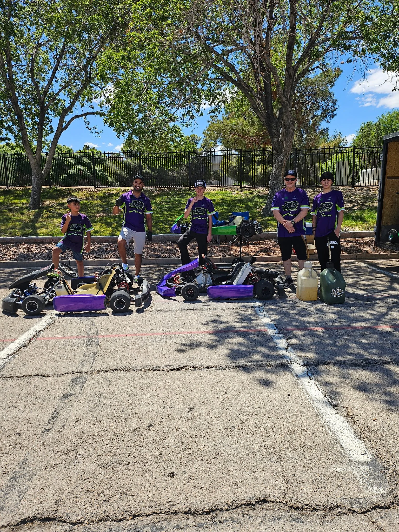 Group of five kids and adults standing outdoors in a parking lot, next to go-karts, with a large tree and a black fence in the background, sunny day with clear blue sky.